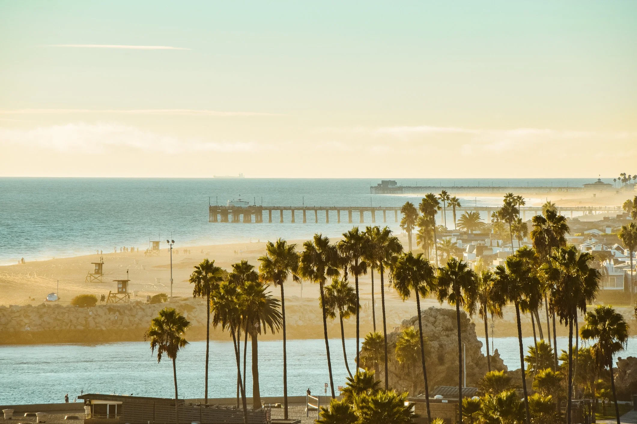 Beach scene with palm trees, sandy shore, pier extending into the ocean, and buildings along the beachfront during sunset