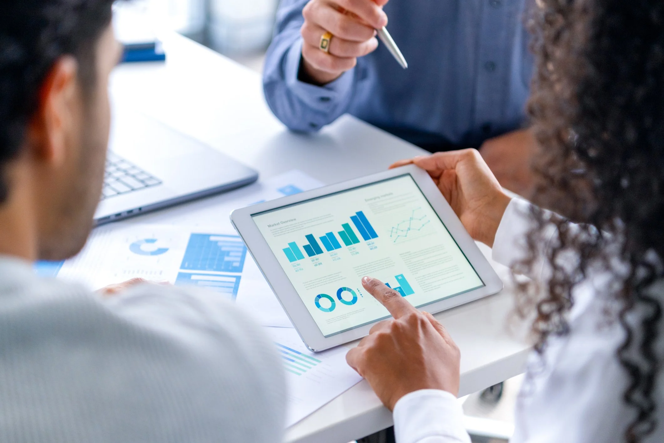 Two people reviewing data and charts on a tablet during a business meeting, with printed graphs and a laptop on the table.