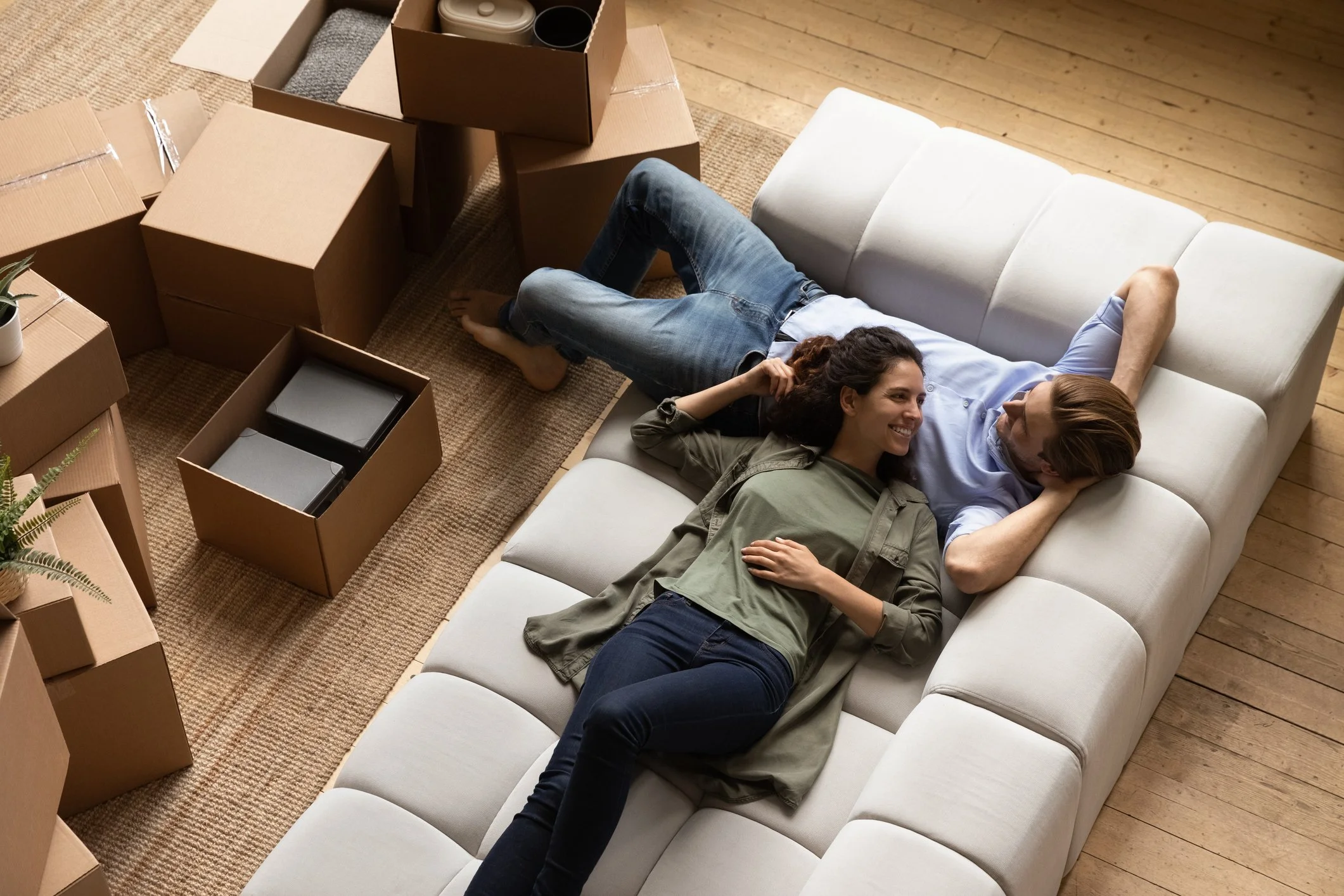 A young man and woman lying on a white sofa, smiling and talking, surrounded by moving boxes, in a room with wooden flooring.