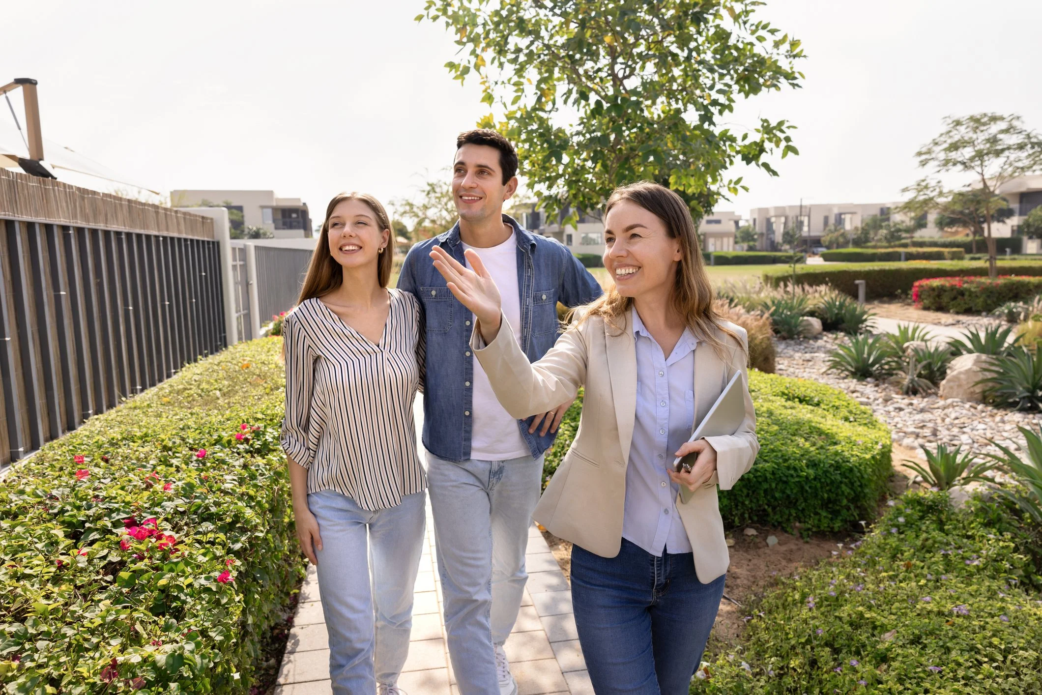 Three people, two women and one man, walking outdoors in a landscaped area with bushes and plants, and smiling while talking.