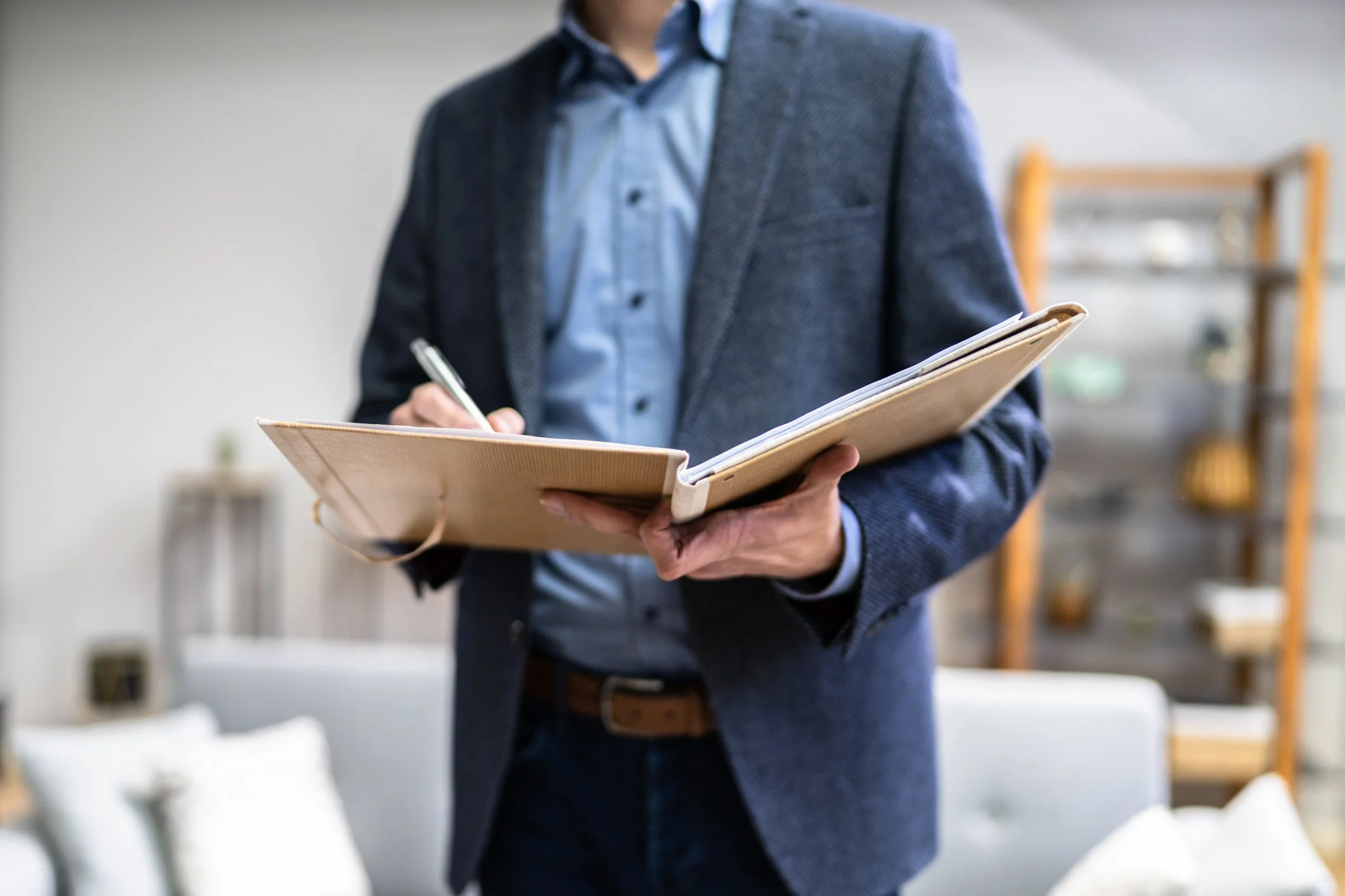 A man in a blue shirt and gray blazer holding an open notebook and pen, standing in an office or living room.