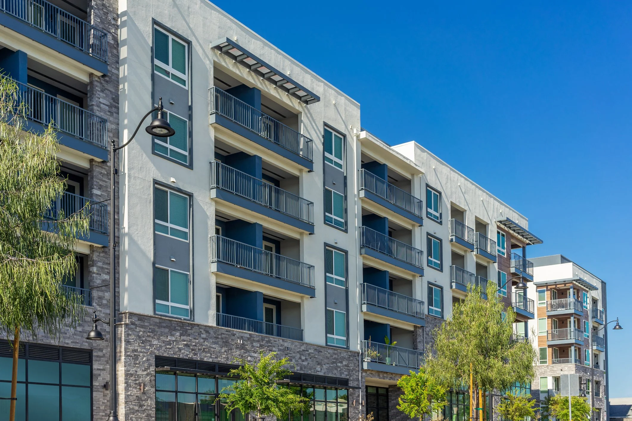Modern multi-story residential buildings with balconies, large windows, and street lamps in the foreground under a clear blue sky.