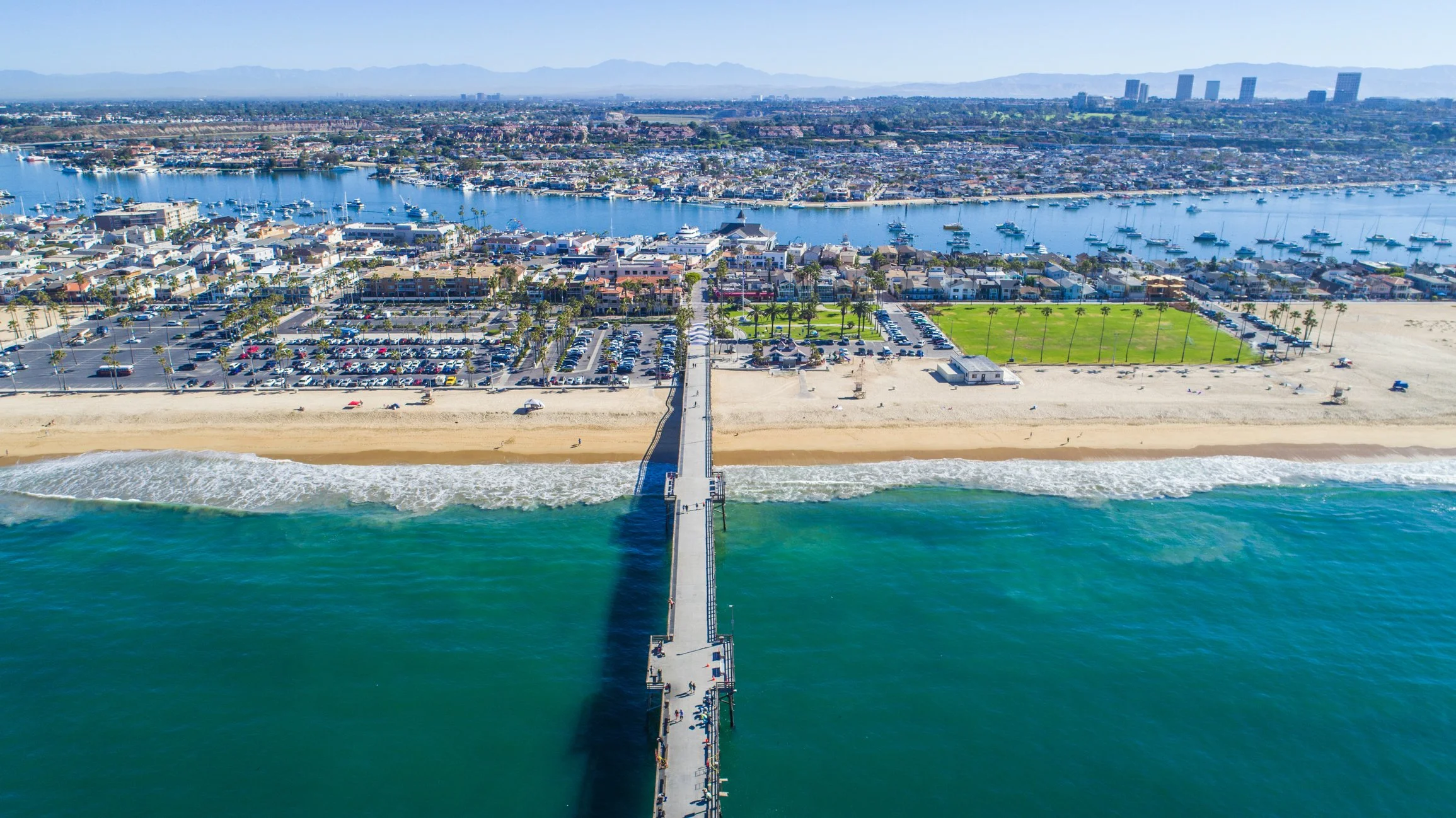 Aerial view of a beach with a pier leading into the ocean, a parking lot, and a cityscape across a waterway with boats.