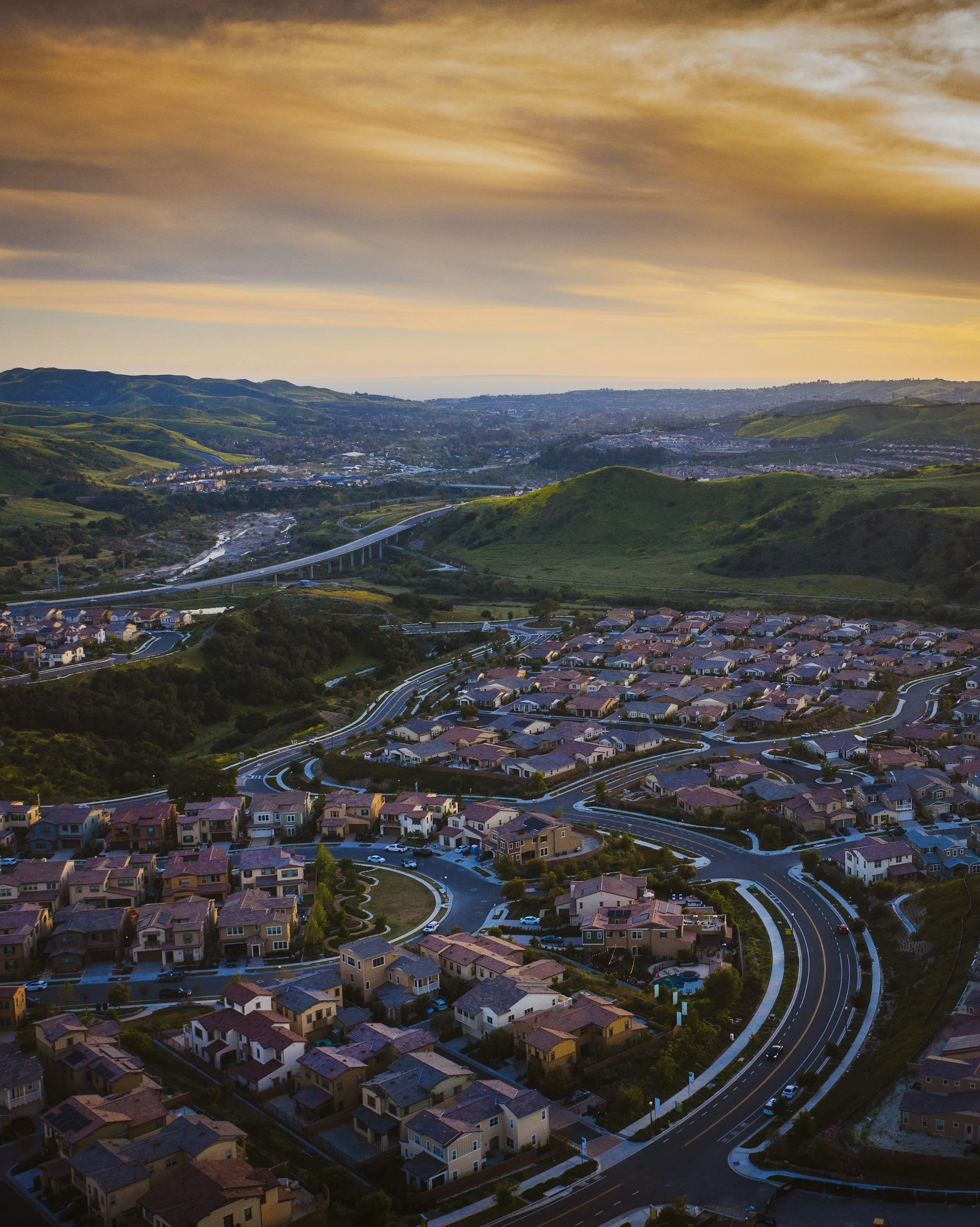 Aerial view of a suburban neighborhood with winding roads, houses with tiled roofs, and green hills at sunset.