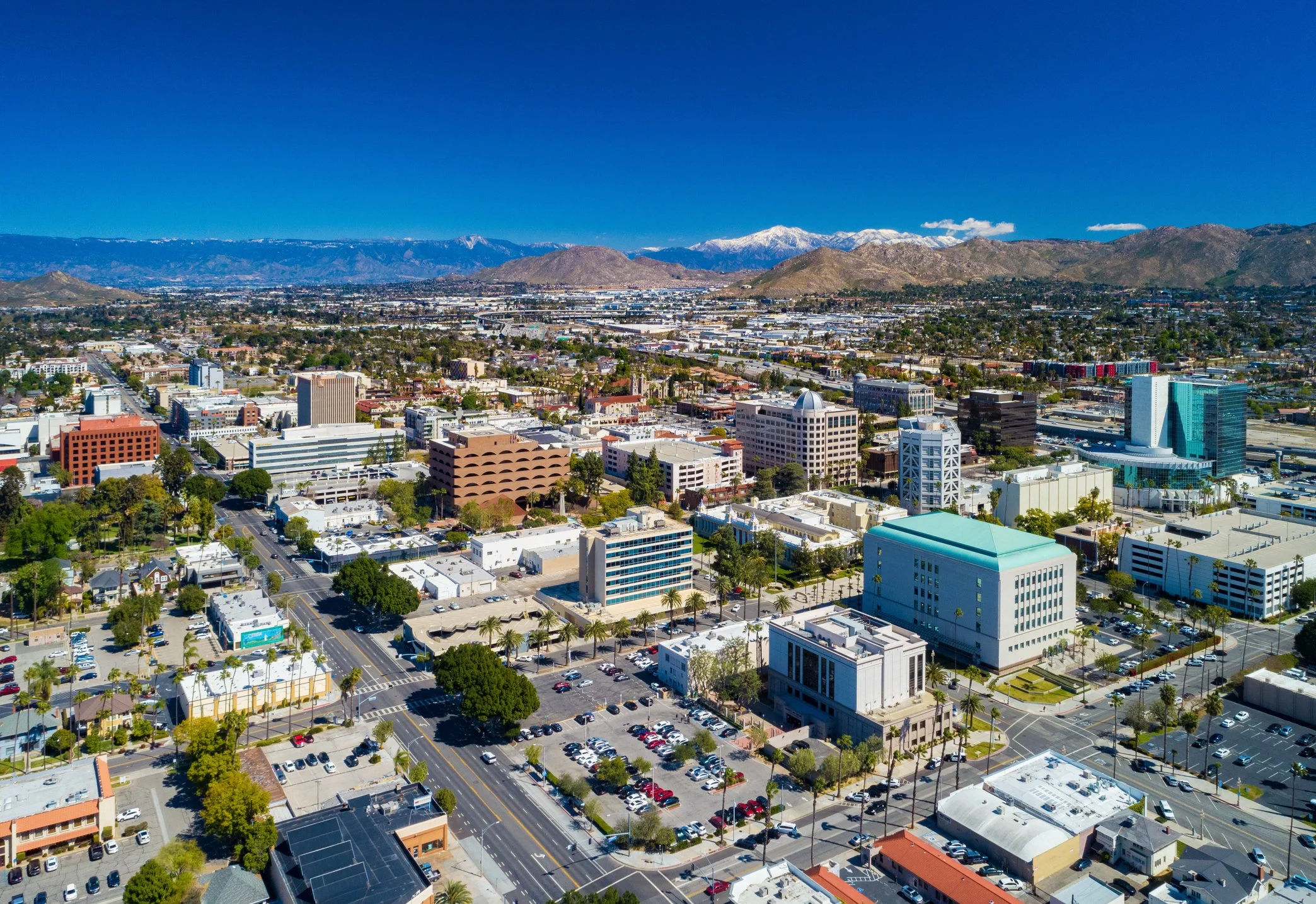 Aerial view of a city with modern buildings, green trees, parking lots, and mountains in the background under a clear blue sky.