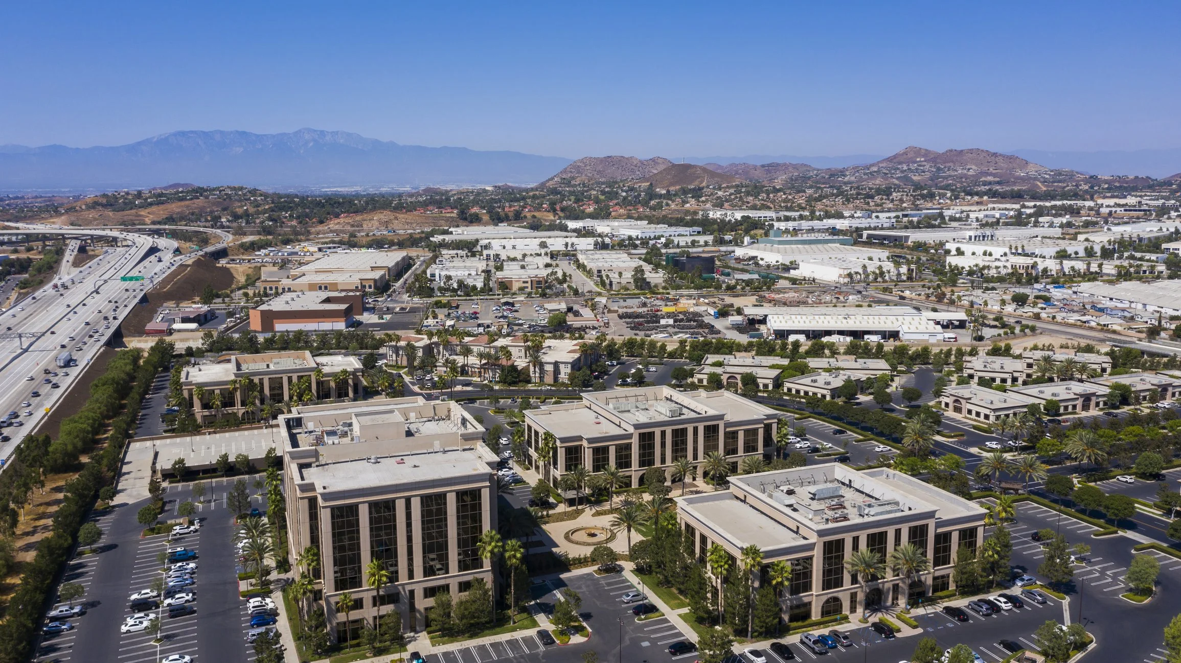 Aerial view of a commercial and office complex with parking lots, surrounded by industrial buildings and highways, with mountains in the background.