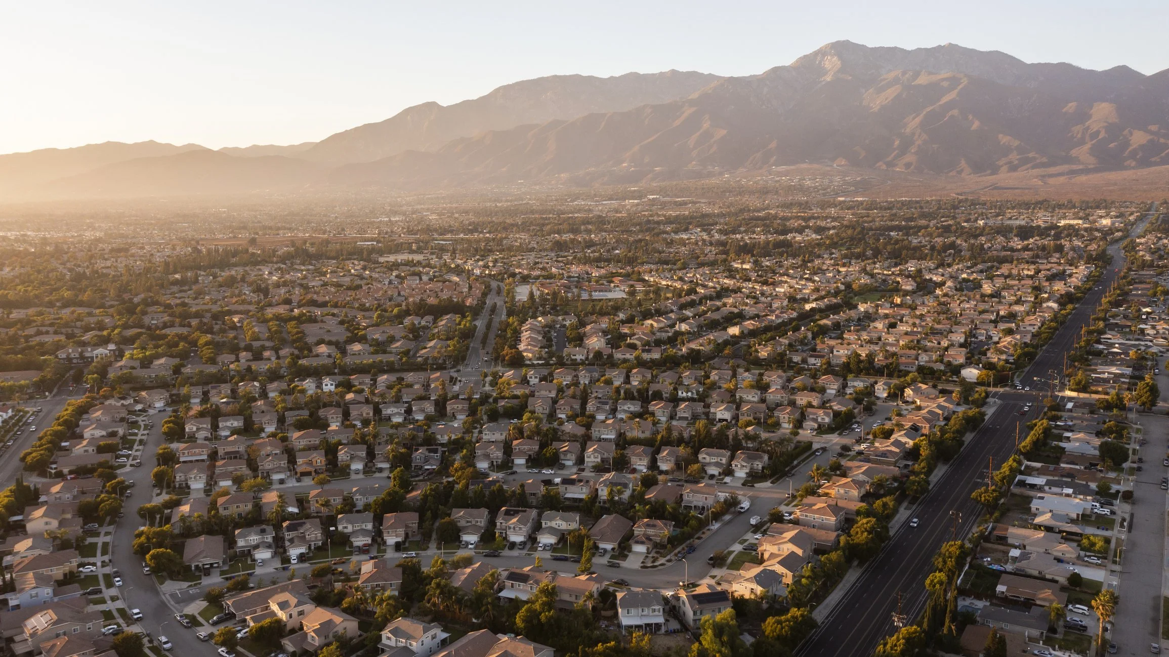 An aerial view of a suburban neighborhood at sunset with houses, trees, and a main road leading towards mountains in the distance.