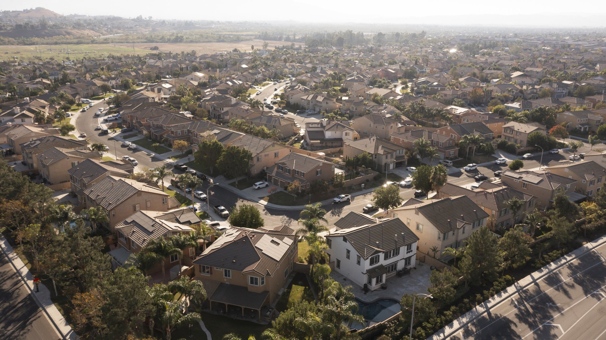 Aerial view of a suburban neighborhood with numerous houses, roads, cars, trees, and a swimming pool in the backyard.