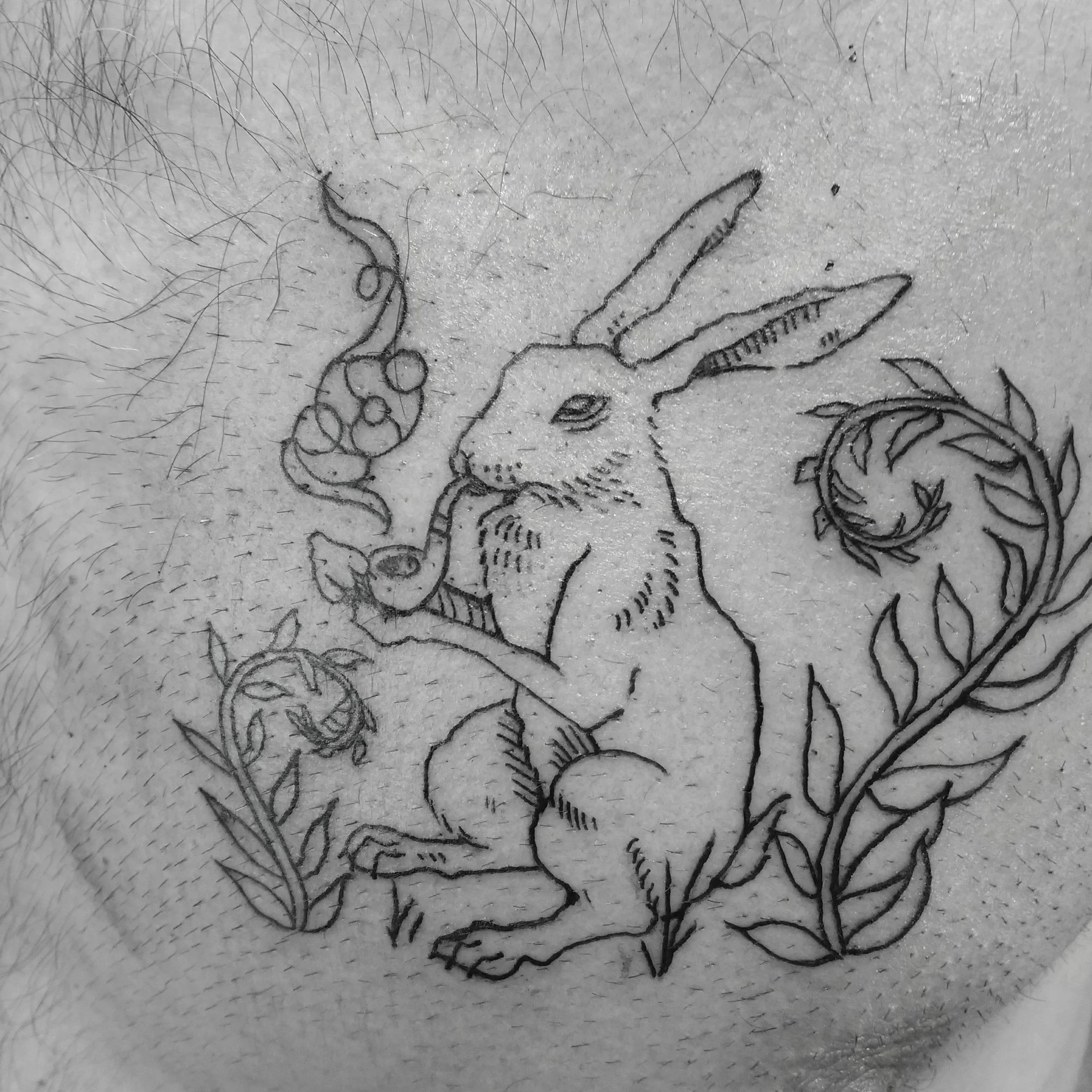 Tattoo of a rabbit with long ears, holding a pipe in its mouth, surrounded by leafy plants.