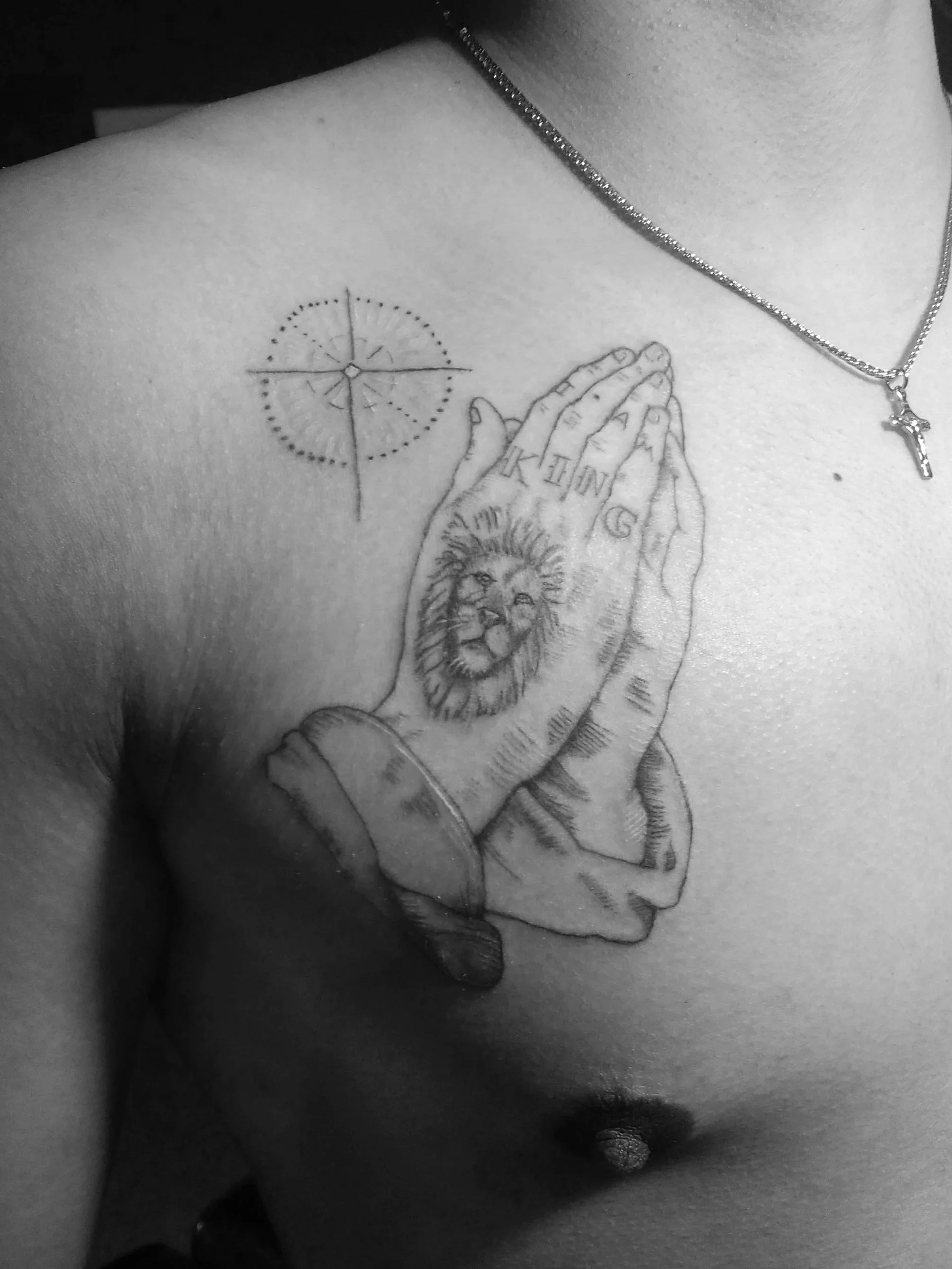 Tattoo of praying hands with a lion's face on one hand and the text 'King' on fingers, with a compass rose above on a person's chest.
