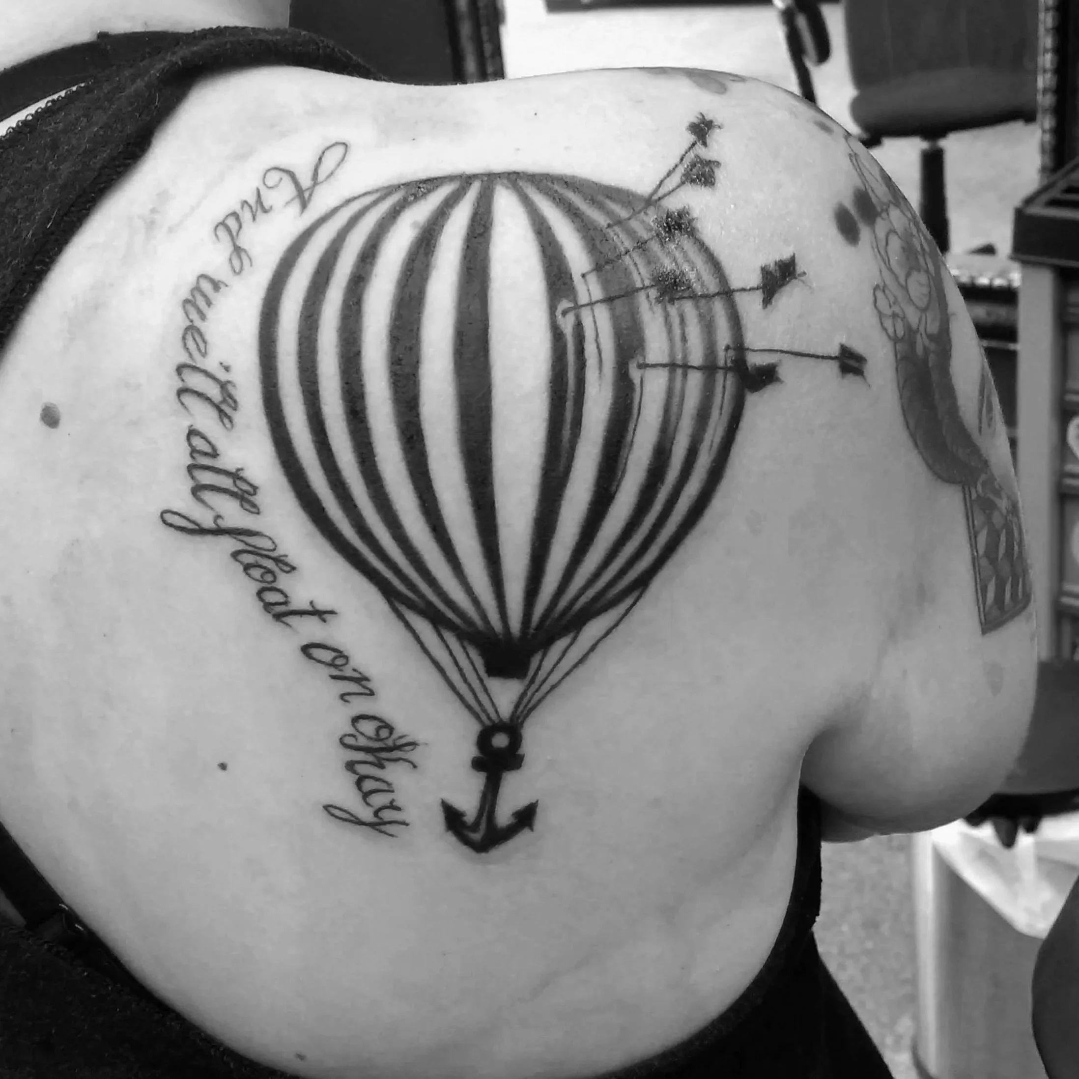 Black and white tattoo of a hot air balloon with an anchor hanging from it on a person's shoulder and upper back, with some decorative text around.