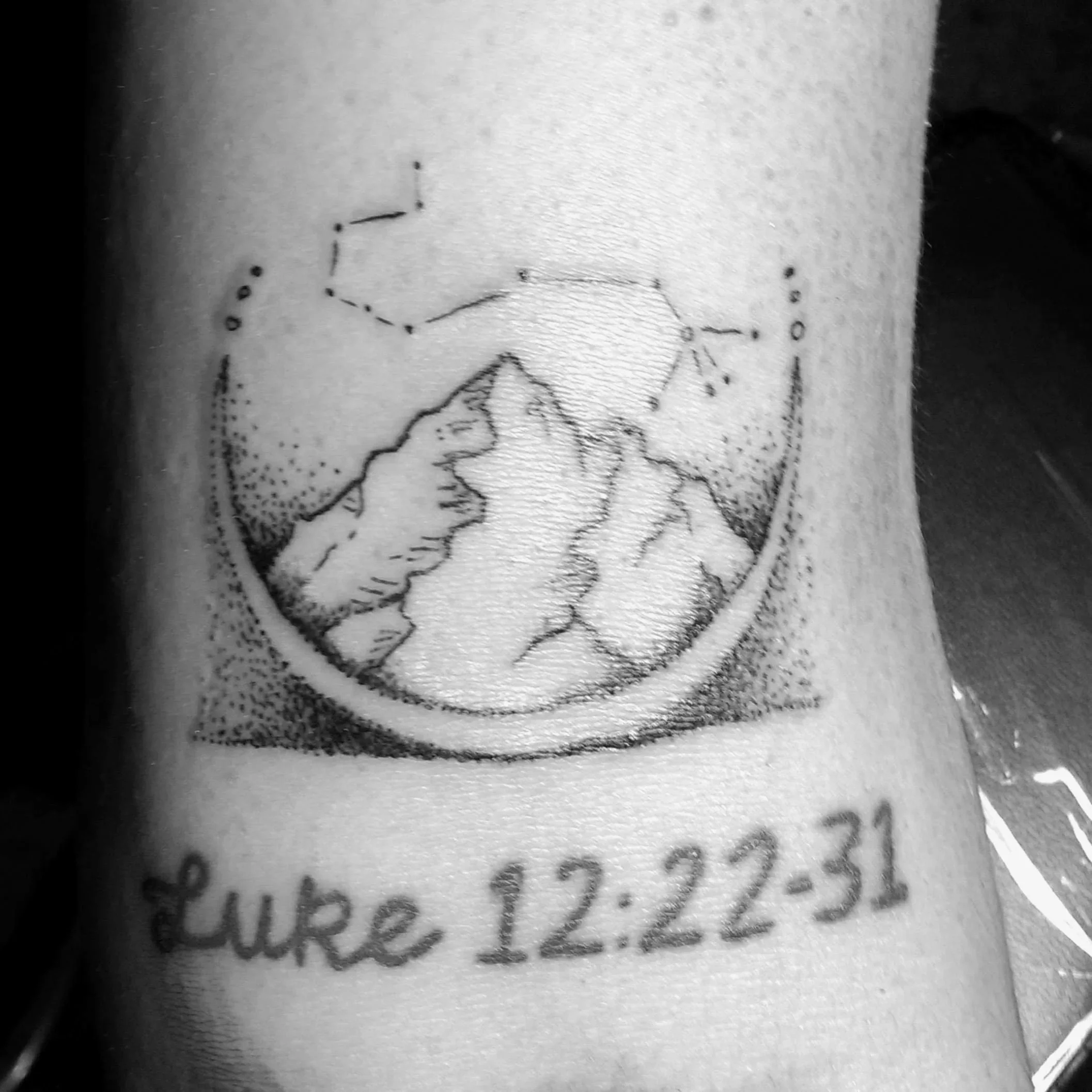 Tattoo of a mountain landscape with stars and constellations above, and the date 'Luke 12:22-31' written below.