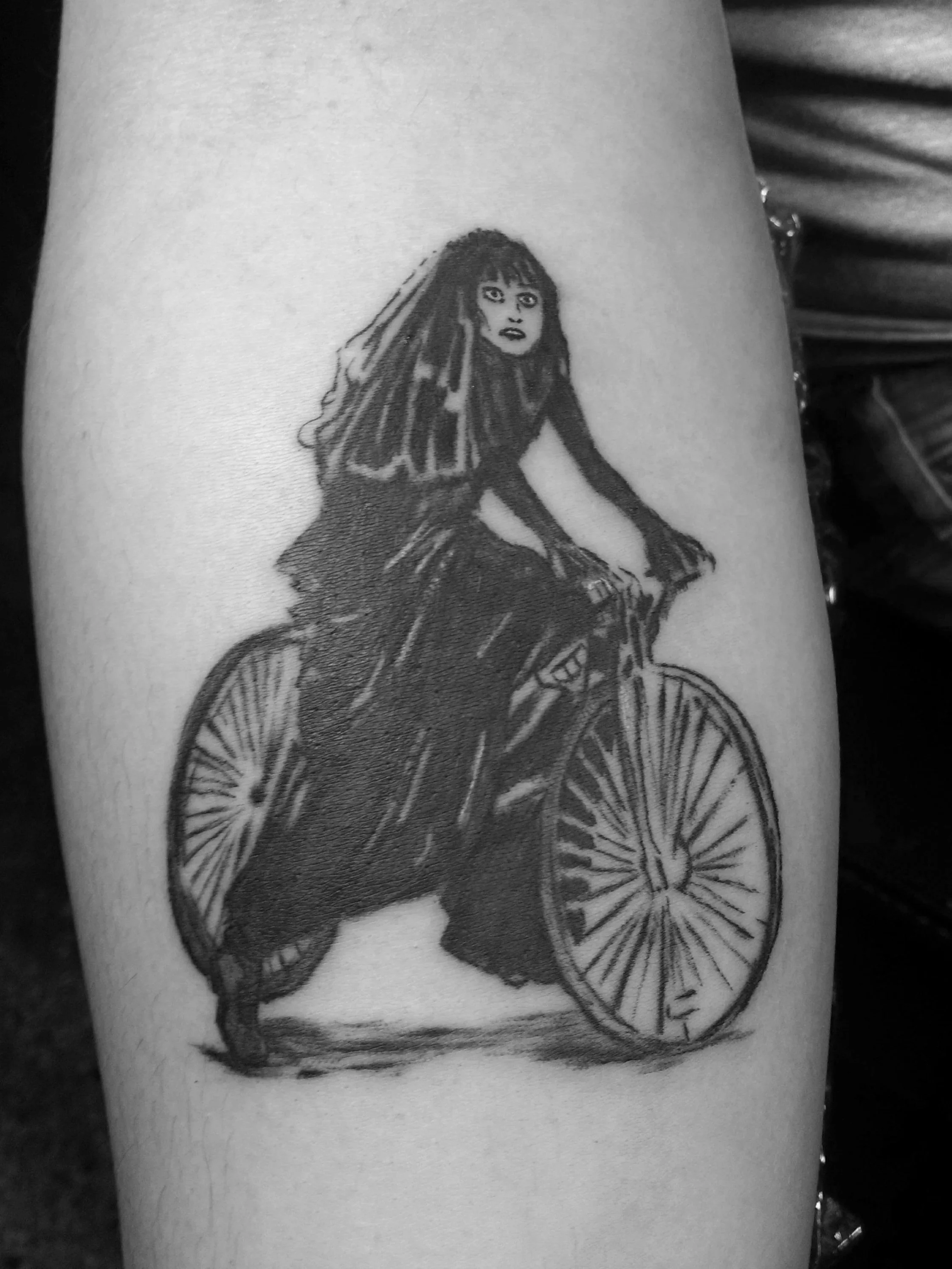 Black and gray tattoo of a woman with long dark hair riding a vintage bike with large wheels.