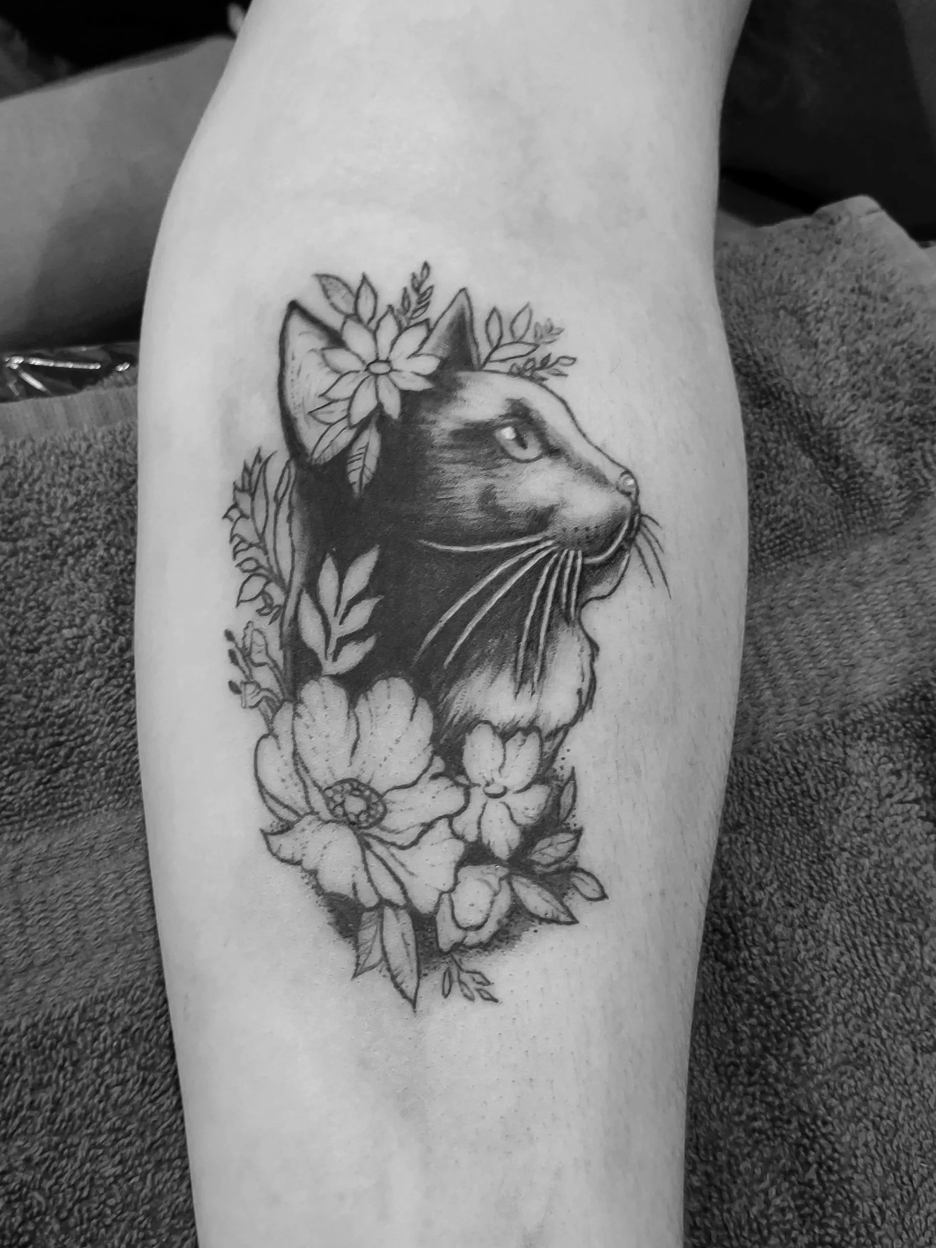 Black and gray tattoo of a cat's profile surrounded by flowers on a person's arm.