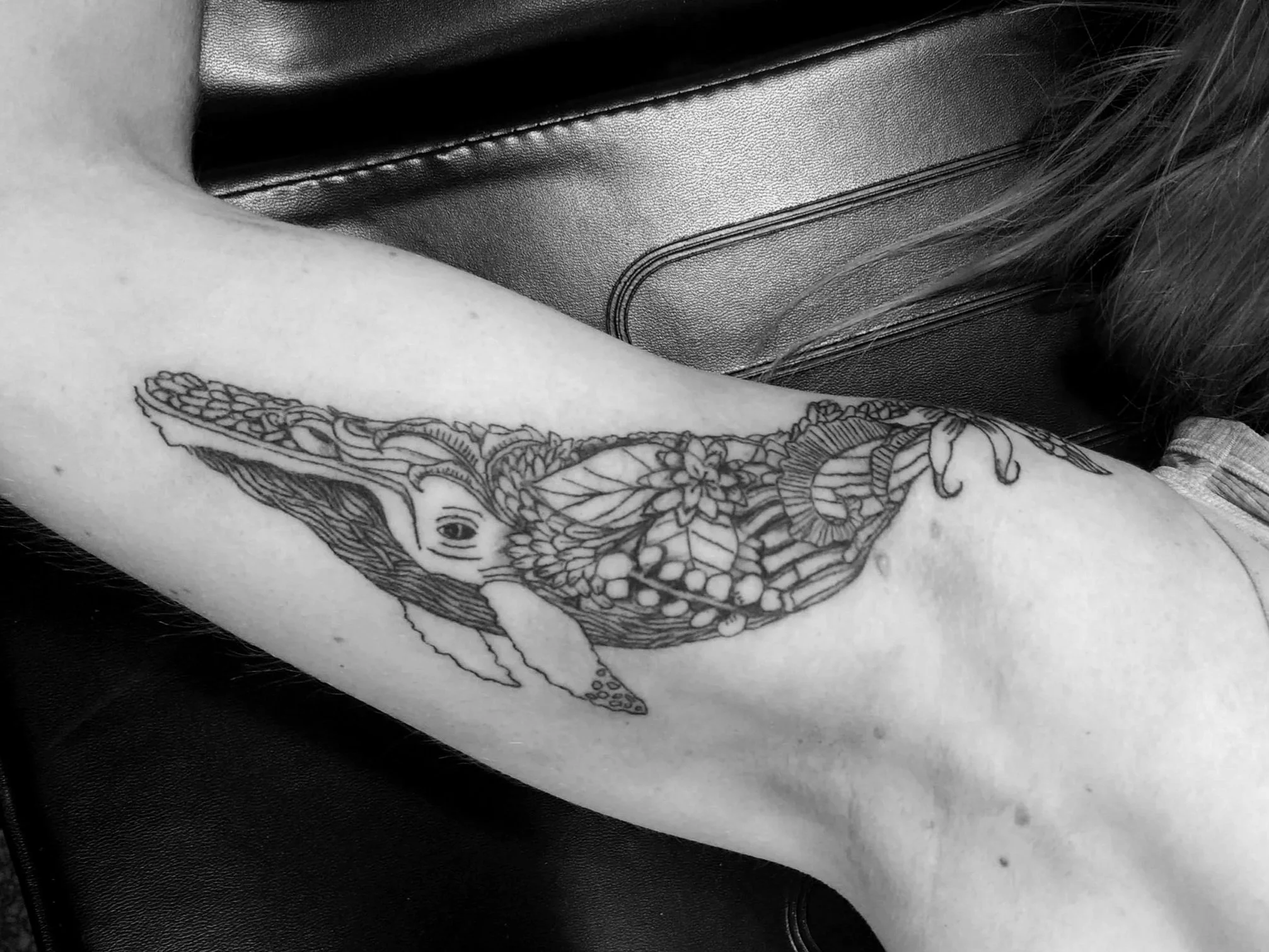 Black and white photo of a tattoo of a whale with floral and botanical designs on a person's arm.