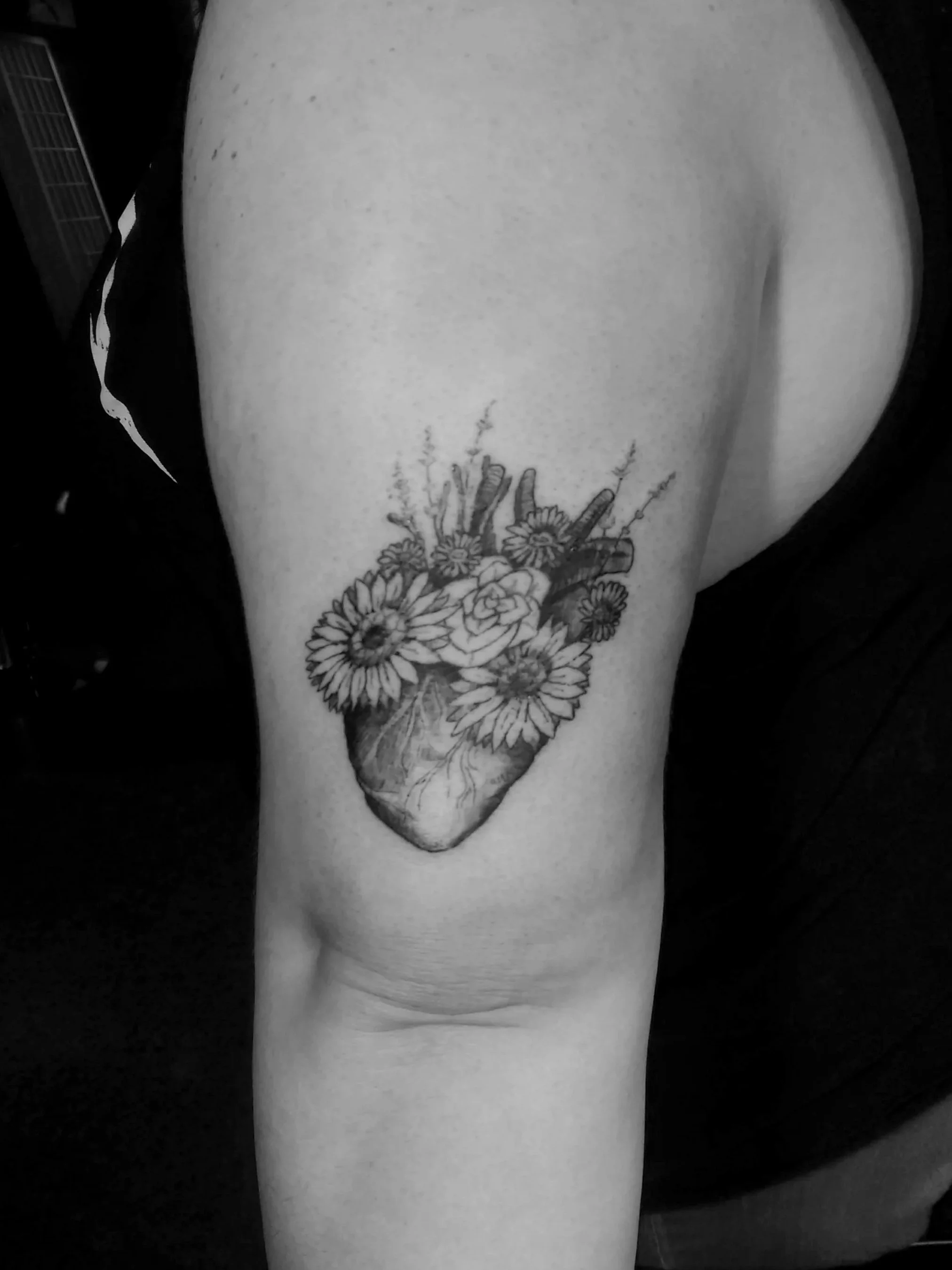 Black and white tattoo of a human heart with flowers and plants emerging from it, located on the upper arm.