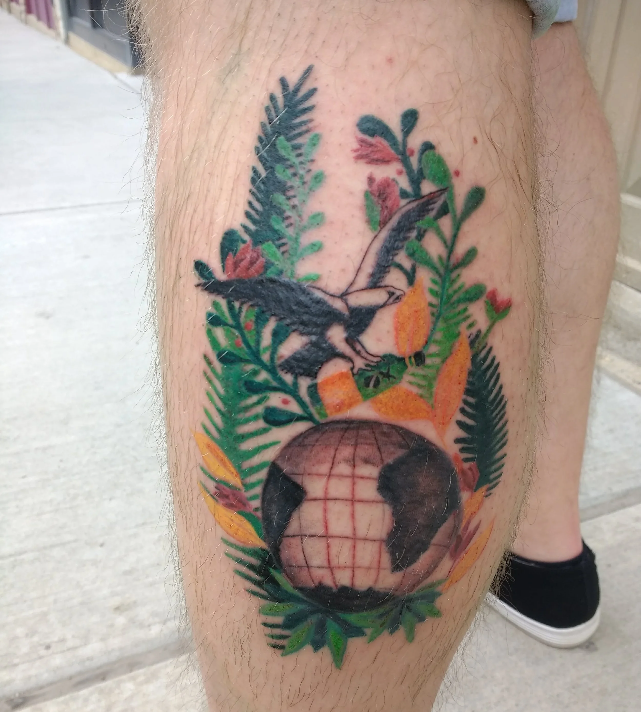 A colorful tattoo featuring a bird in flight surrounded by green leaves and orange flowers, with a globe at the bottom.
