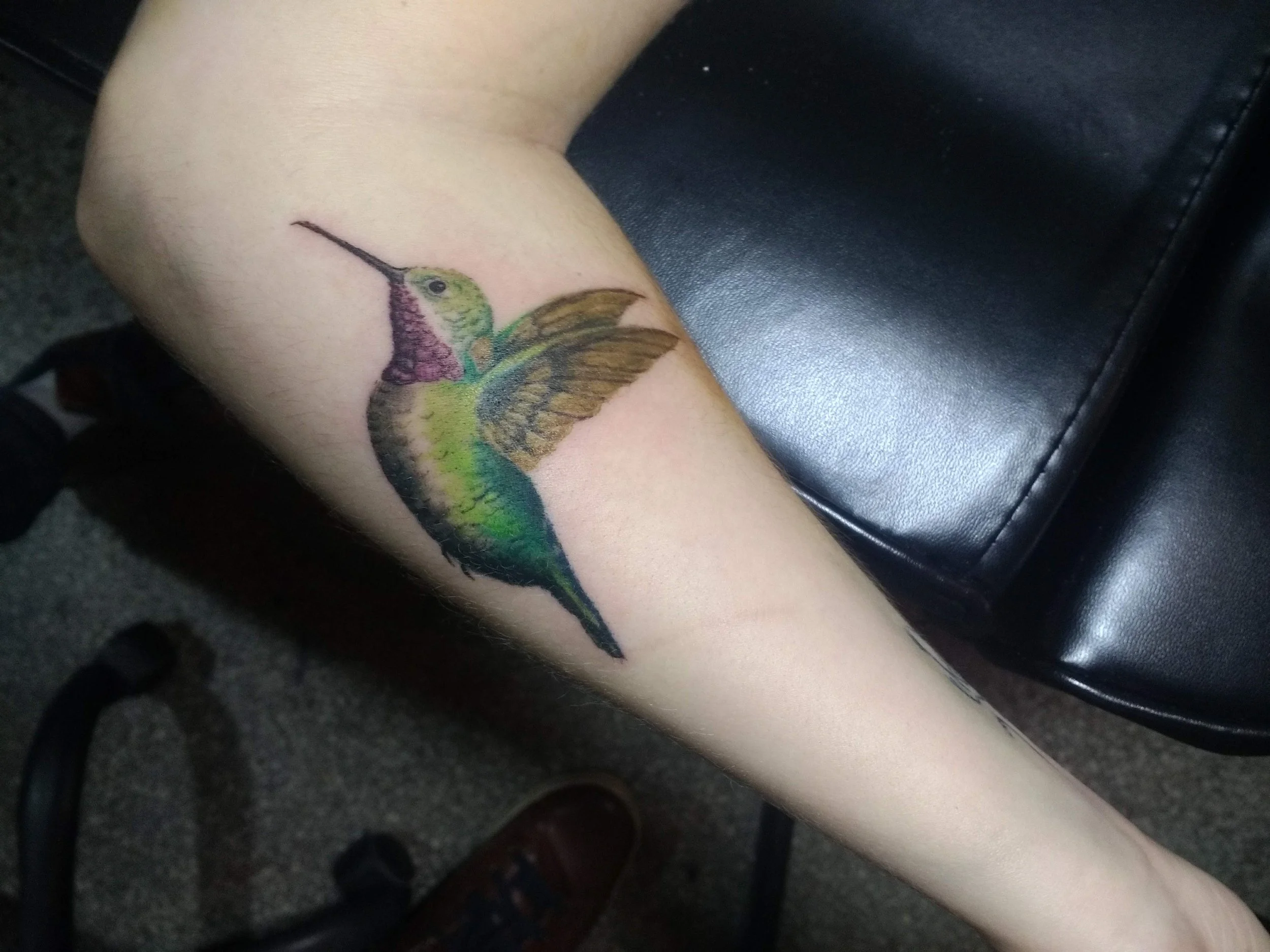 Colorful hummingbird tattoo on forearm, depicting the bird with green, purple, yellow, blue, and black feathers, perched on a surface.