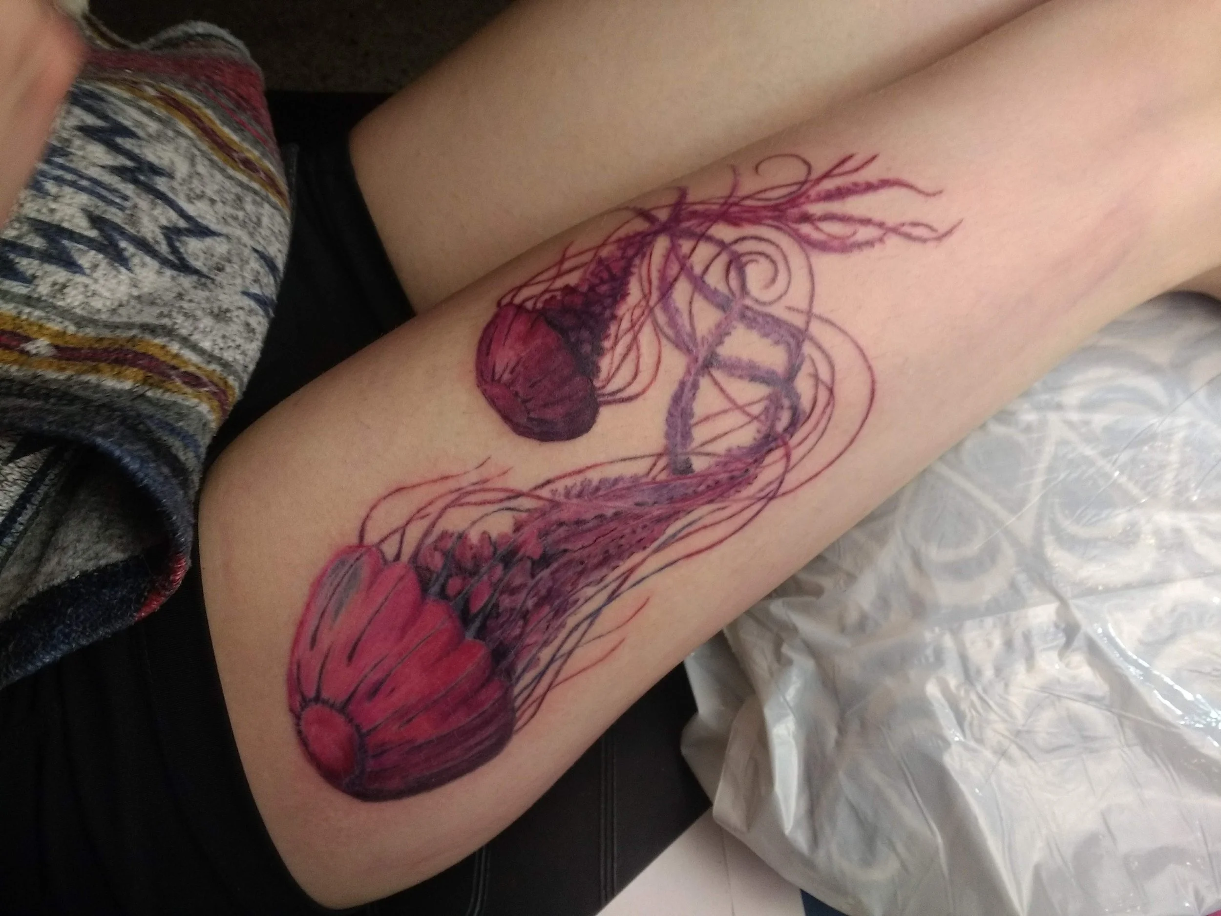 A fresh jellyfish tattoo in progress on a person's thigh, showing detailed outlines and colored sections of the jellyfish's bell and tentacles.