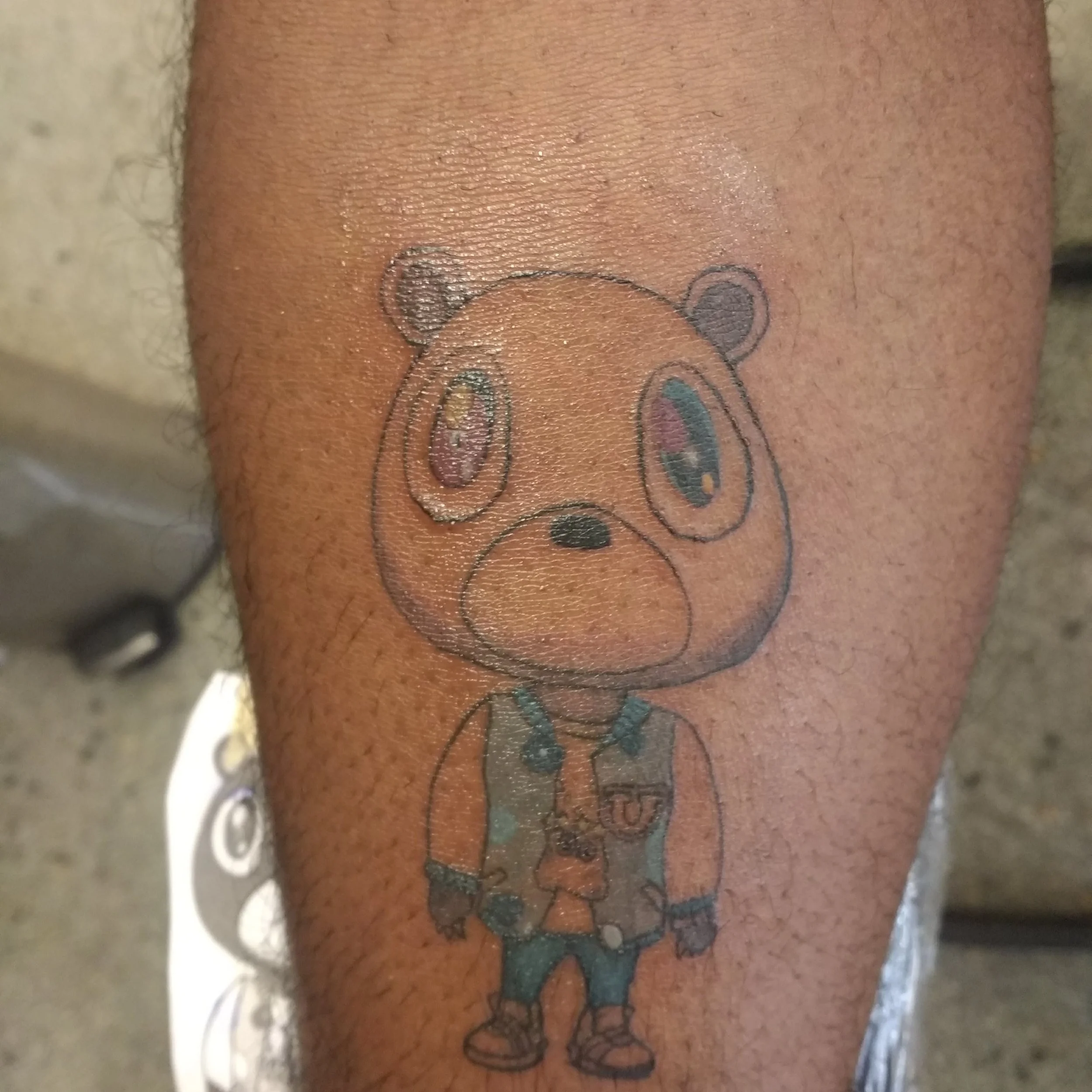 Tattoo of an animated bear character with big eyes, wearing a school uniform with a letter 'U' on the jacket, on a person's leg.