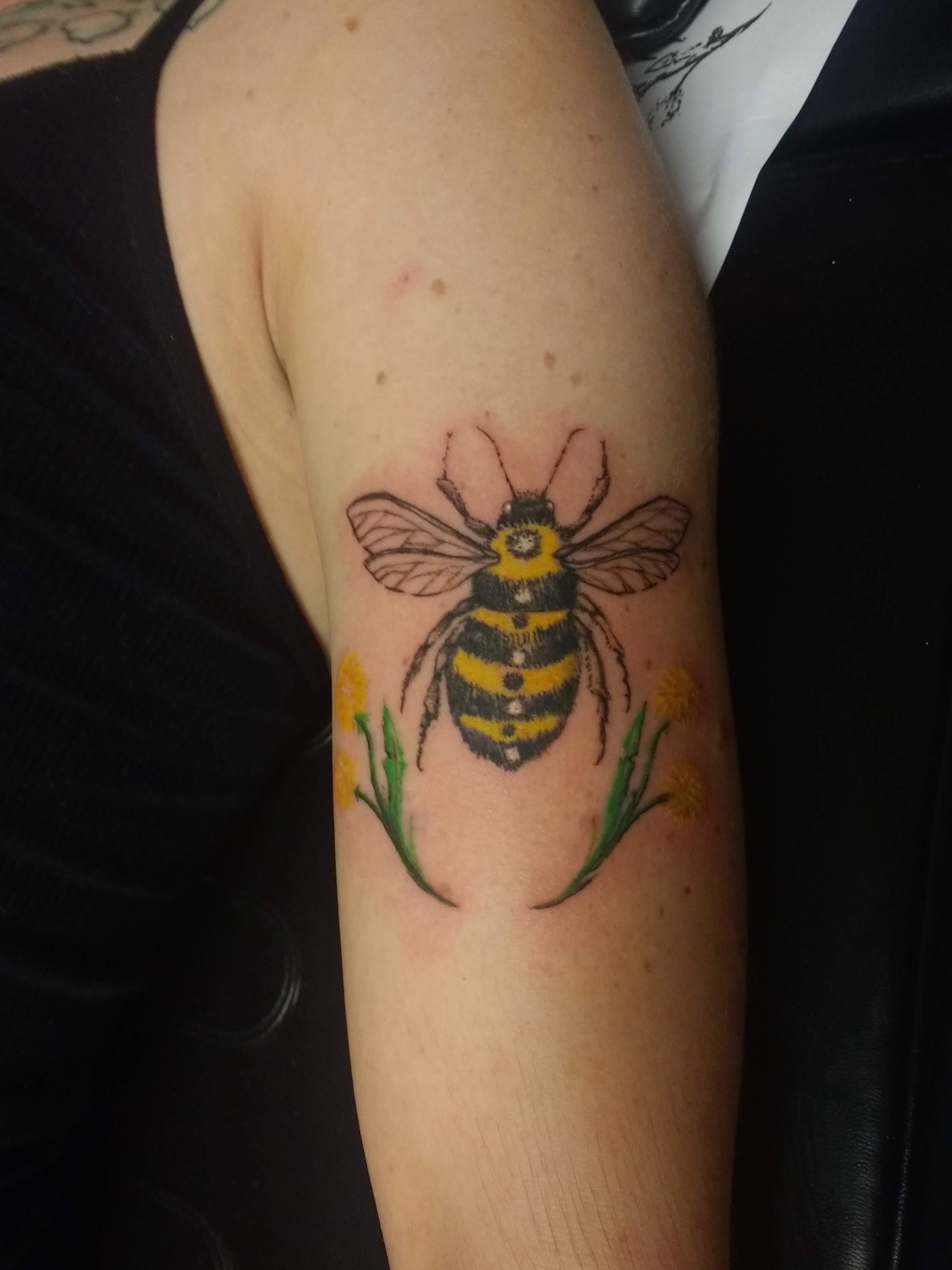Tattoo of a bee on a person's thigh, with yellow and black stripes, wings, and flowers underneath.
