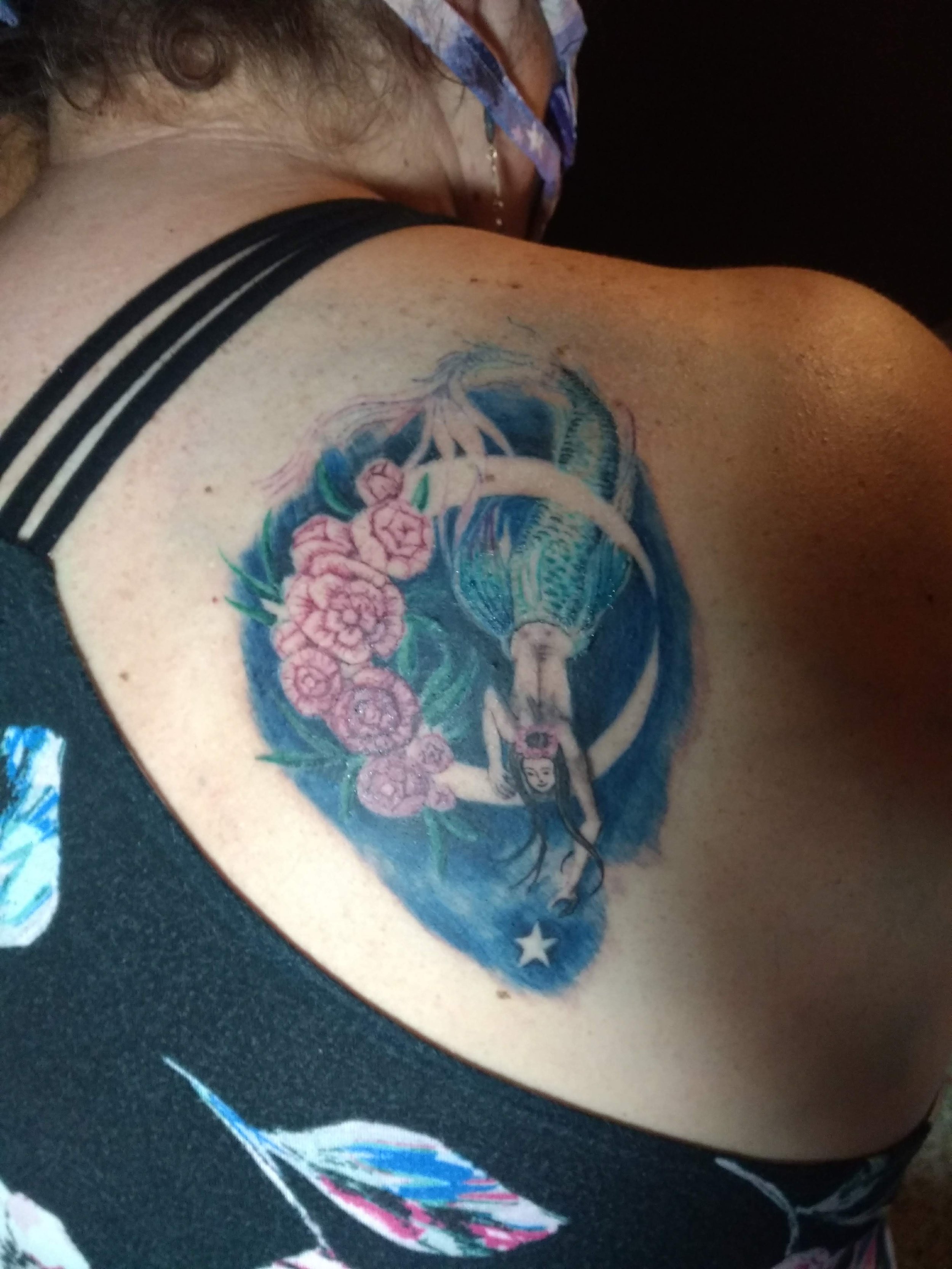 Colorful tattoo of a mermaid with pink flowers on her tail, surrounded by water and a star, on a person's shoulder.