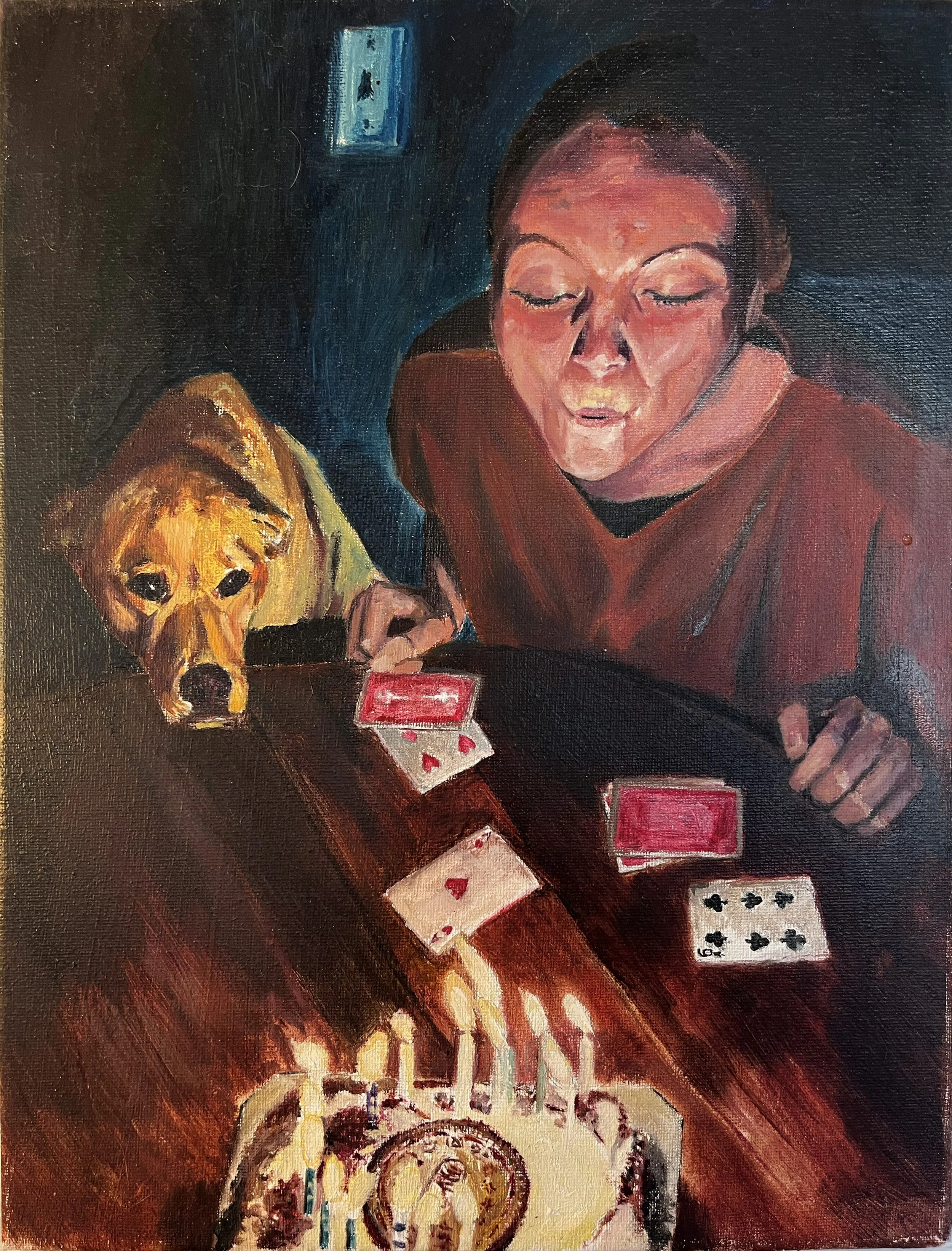 A woman and a dog sitting at a table with a birthday cake and playing cards. The woman is blowing candles out the dog is looking at the cake.