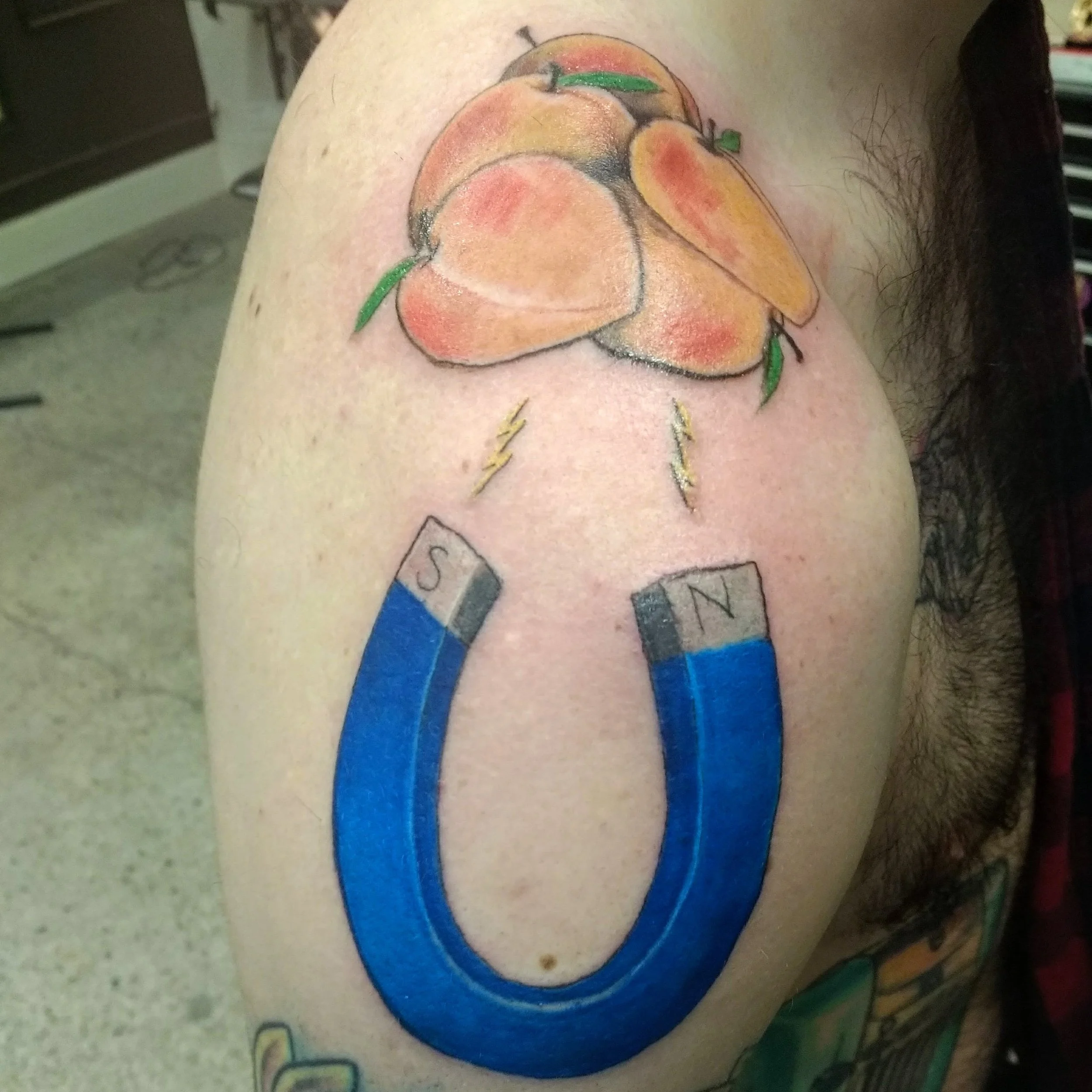 Tattoo of a peach with green leaves above a blue horseshoe magnet with the letters 'S' and 'N' on its ends, and small yellow lightning bolts.