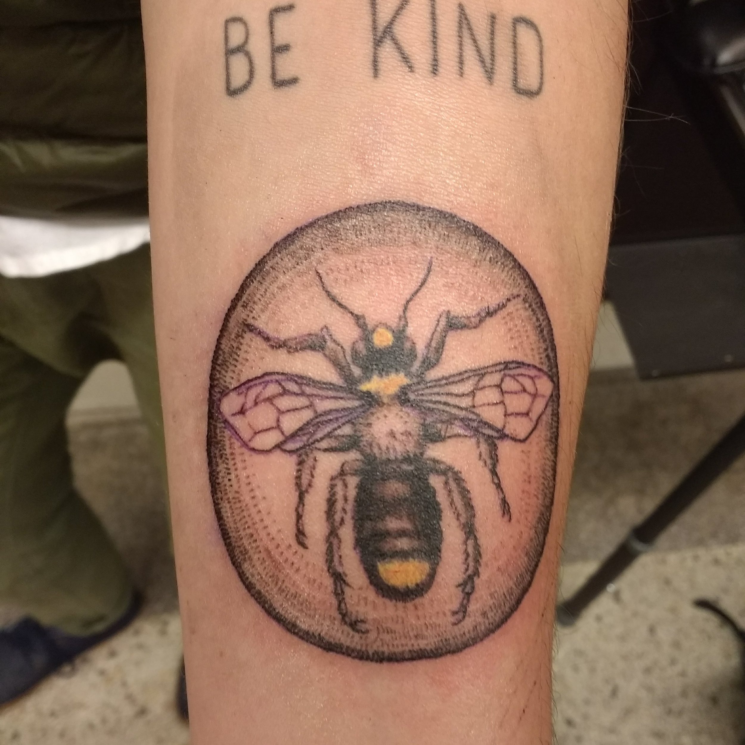 Tattoo of a bee inside a circular design, with the words 'BE KIND' tattooed above it.