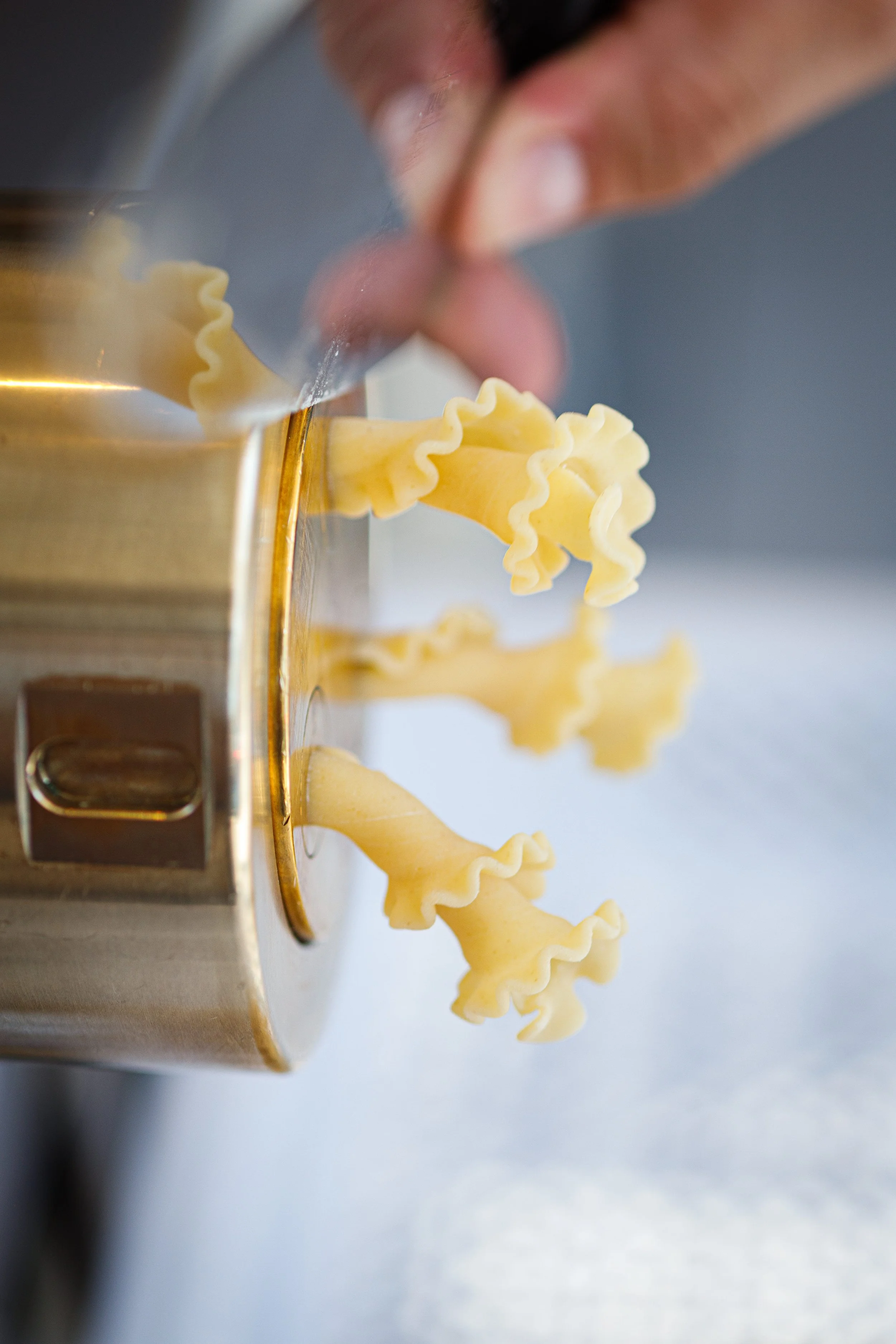 Close-up of a person's hand pushing emptying a pasta maker, with freshly extruded pasta with ruffled edges hanging from the machine.