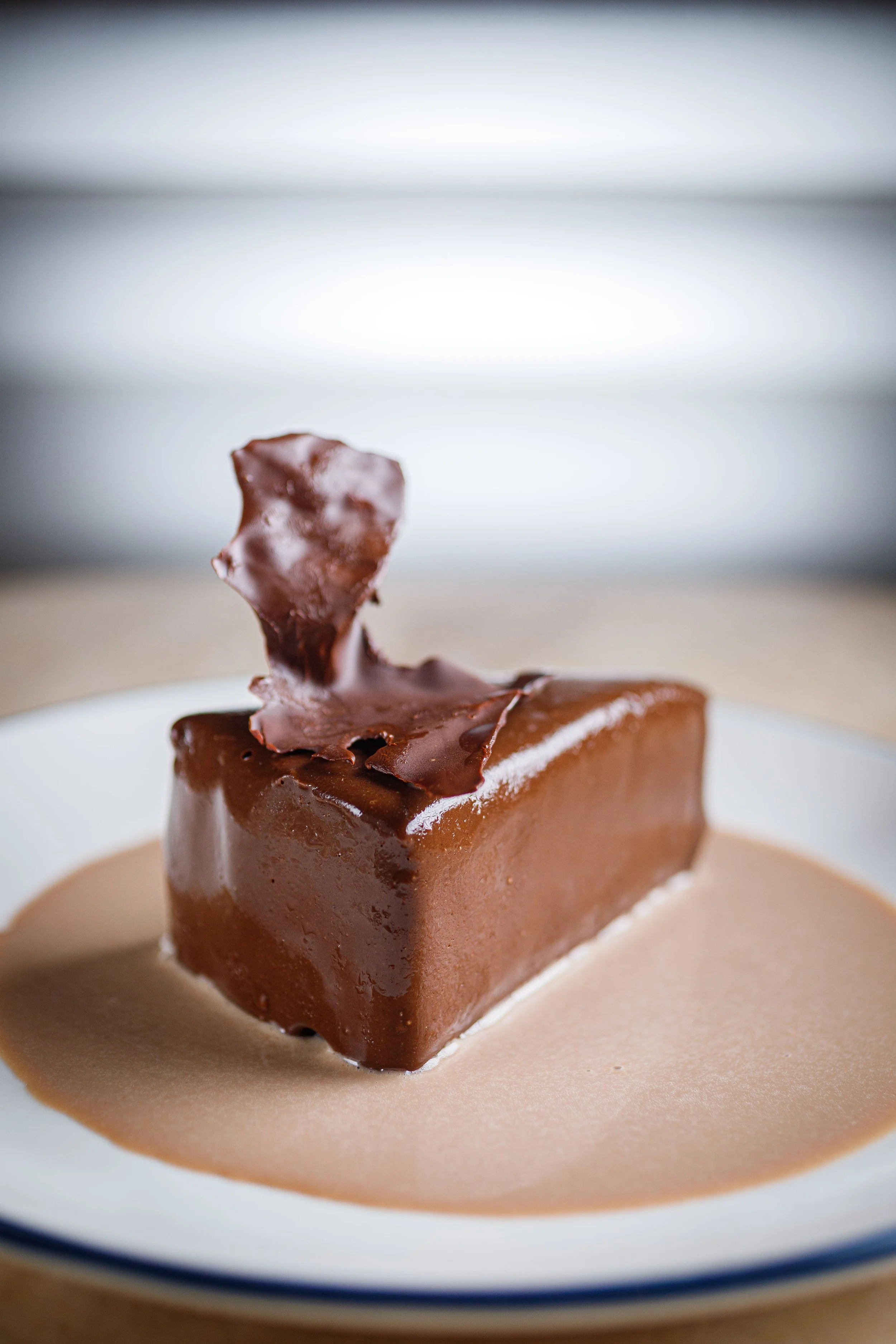 A piece of chocolate dessert with a chocolate piece on top, served in a pool of chocolate sauce on a white plate.