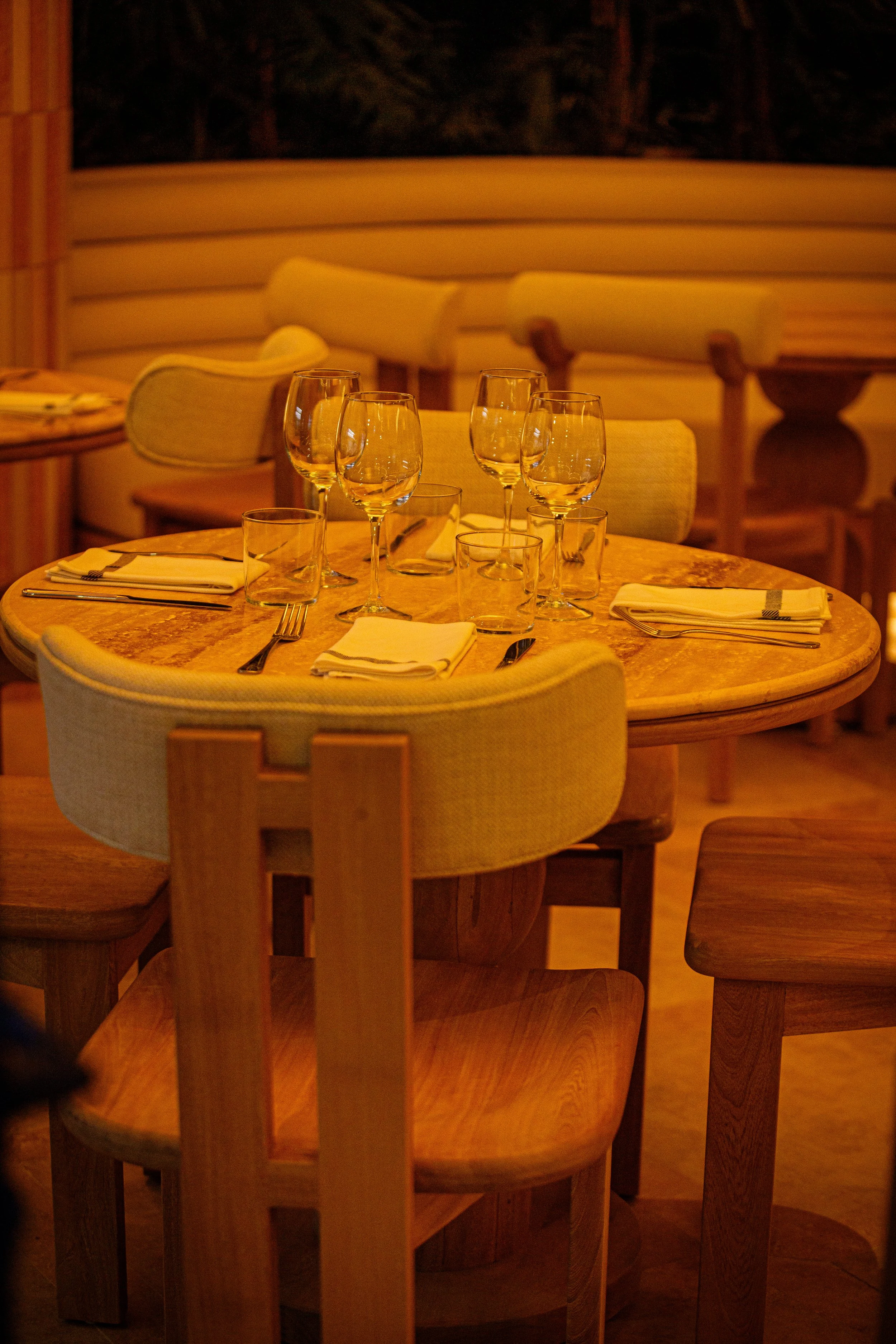 A round wooden dining table set with six wine glasses, six water glasses, napkins, and utensils in a warmly lit restaurant with wooden chairs and decor.