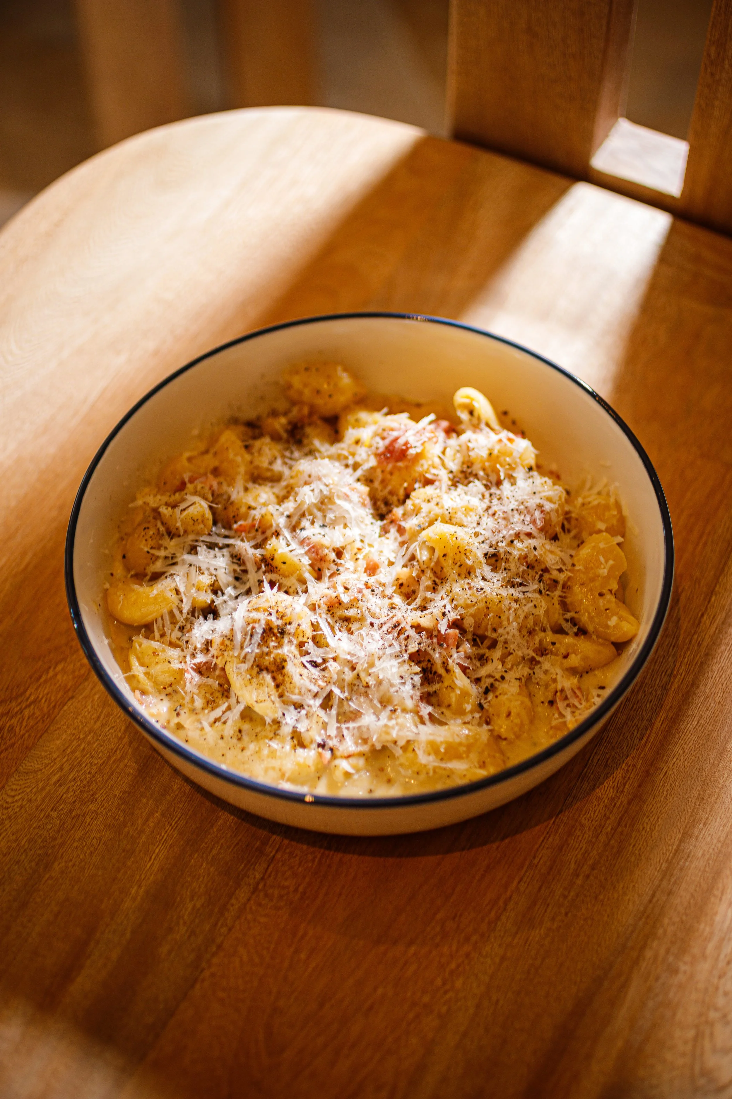 A bowl of creamy pasta with shaved cheese on top sitting on a wooden chair.