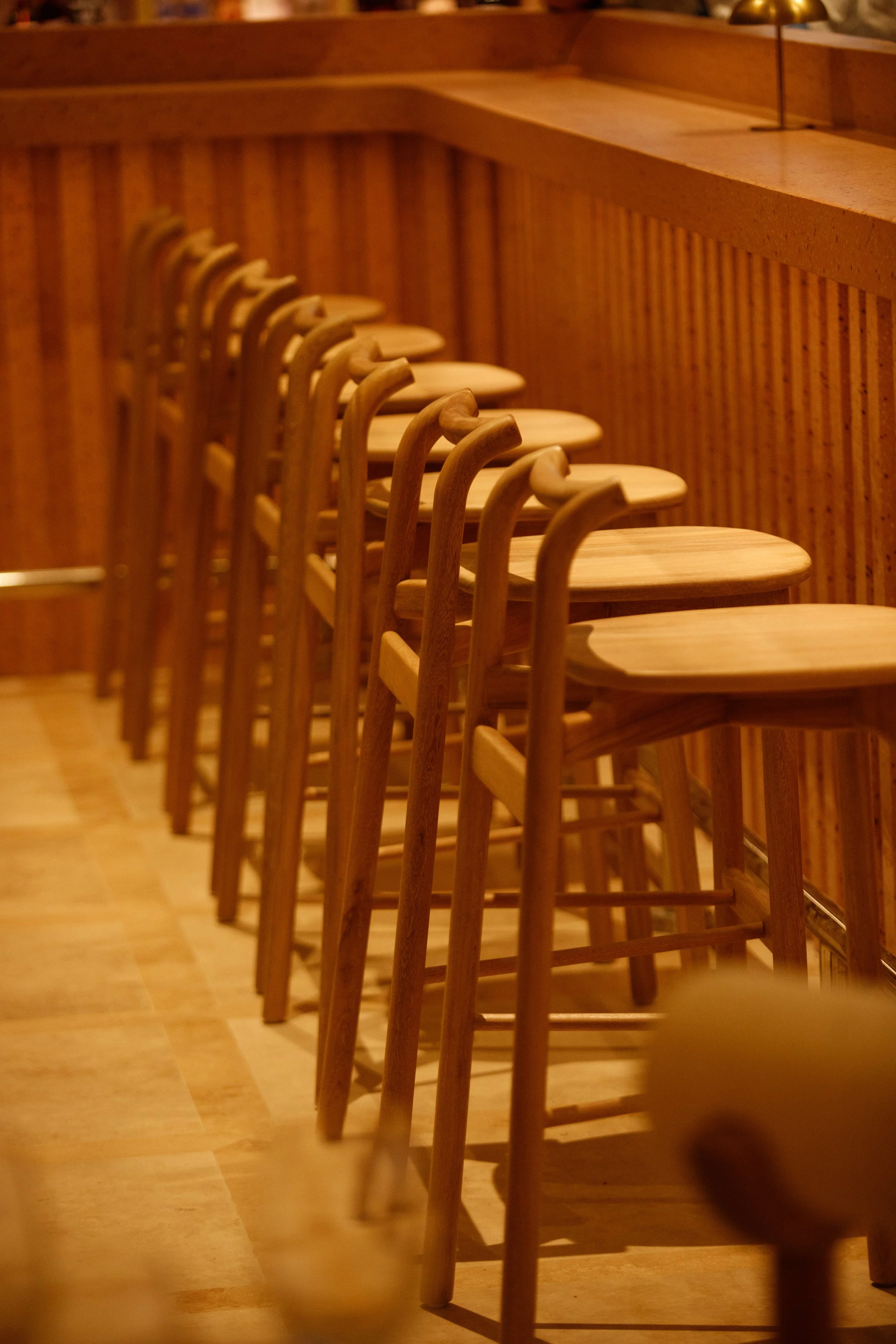 A row of wooden bar stools lined up against a wooden bar counter in a warmly lit setting.
