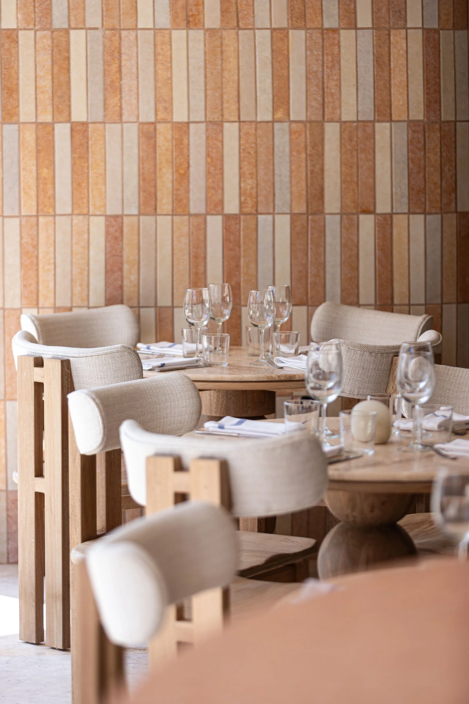 Empty dining room with beige upholstered chairs, round wooden tables set with wine glasses and napkins, against a wall with brick-like patterned tiles at Cuna Puerto Vallarta.