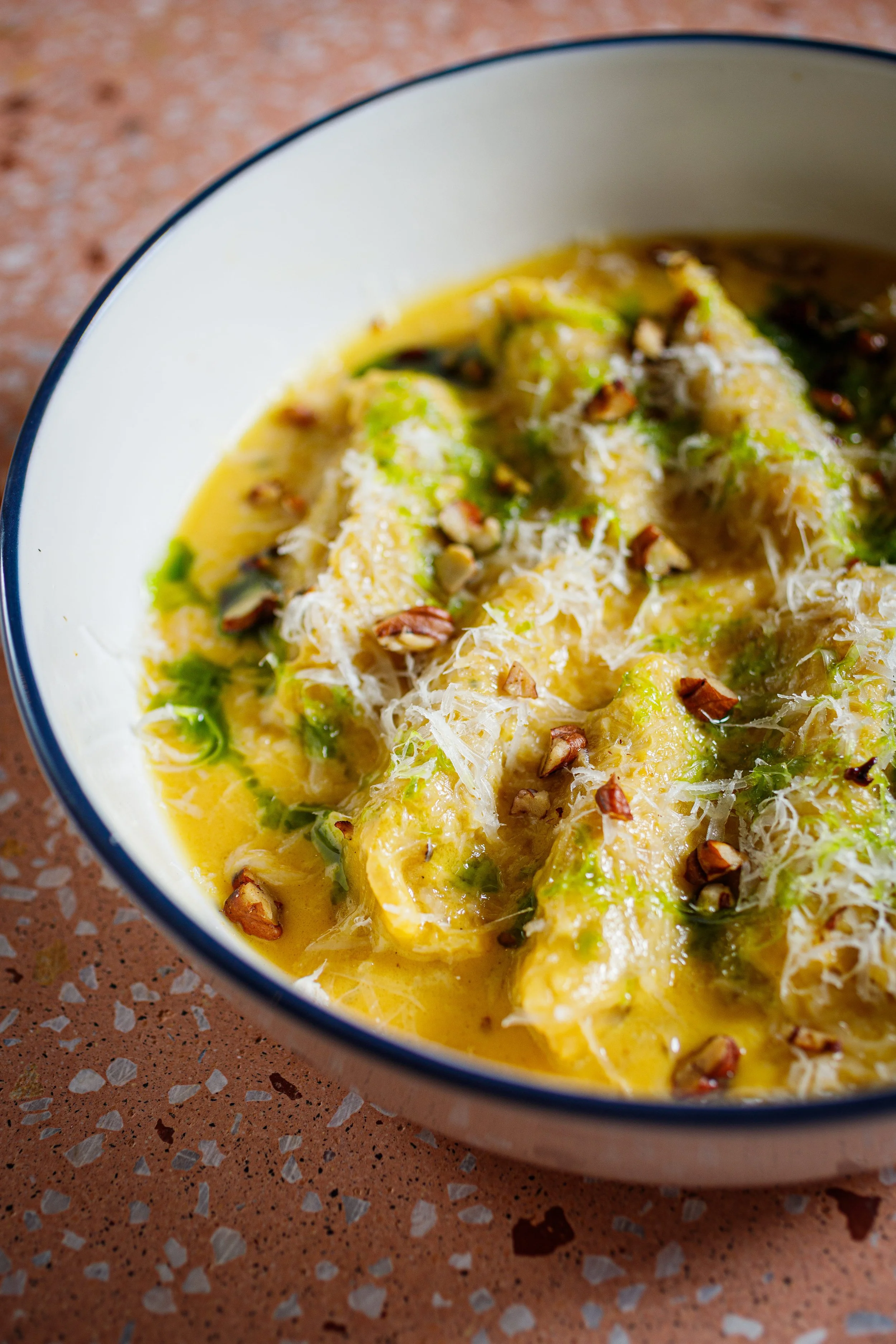 A bowl of cooked zucchini topped with shredded cheese, chopped nuts, and green herbs, sitting on a speckled brown countertop.