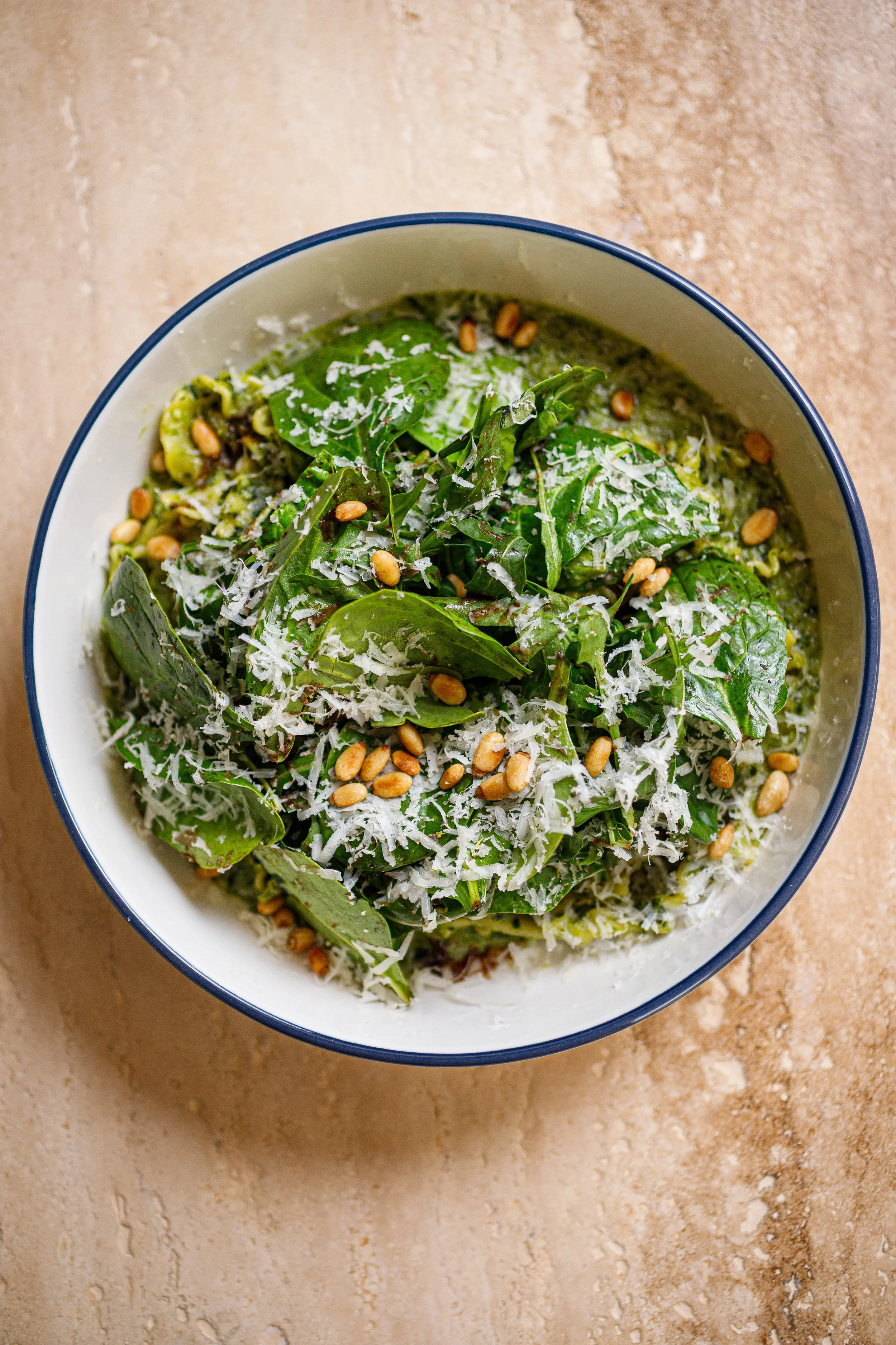 A bowl of green salad topped with shredded cheese and pine nuts on a beige surface.