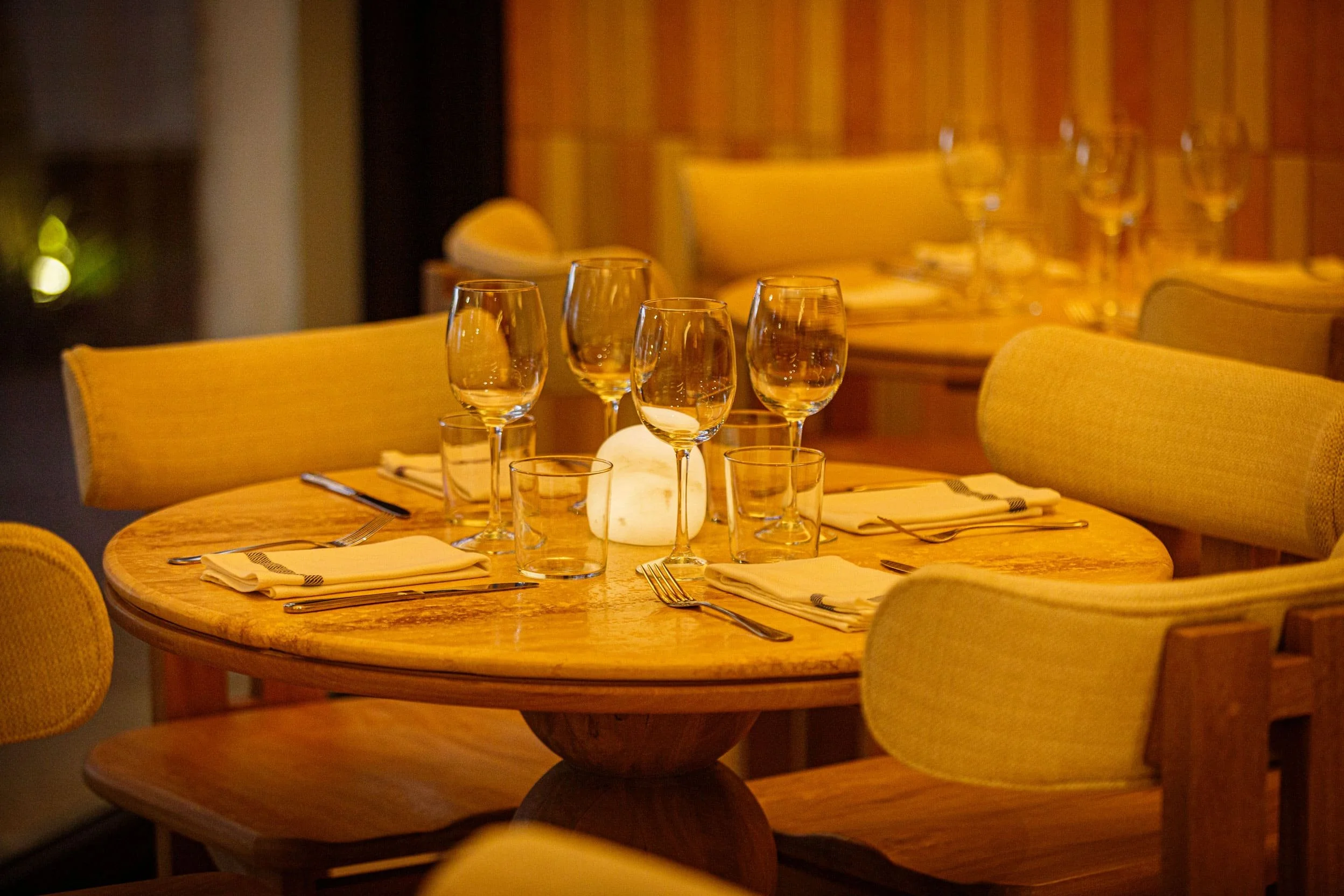 A round wooden dining table set for a meal with four cream-colored cushioned chairs around it, and a centerpiece with a glowing white candle surrounded by empty wine glasses and water glasses, in a warmly lit restaurant.