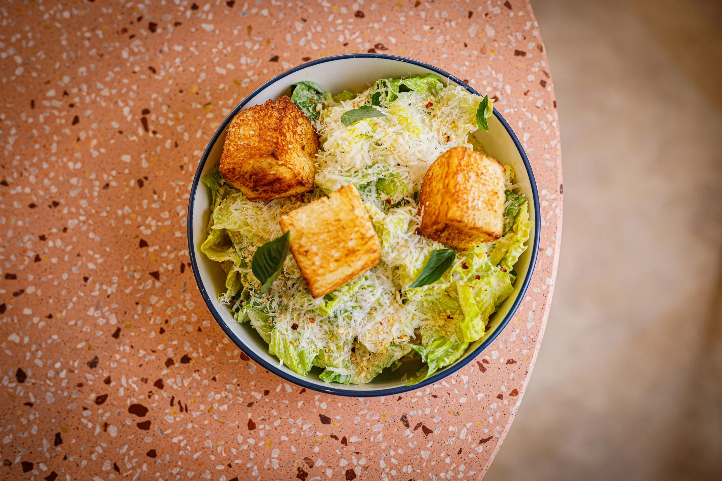 Bowl of Caesar salad with romaine lettuce, grated Parmesan cheese, sliced black pepper, three pieces of crispy croutons, and basil leaves on a pink terrazzo table.
