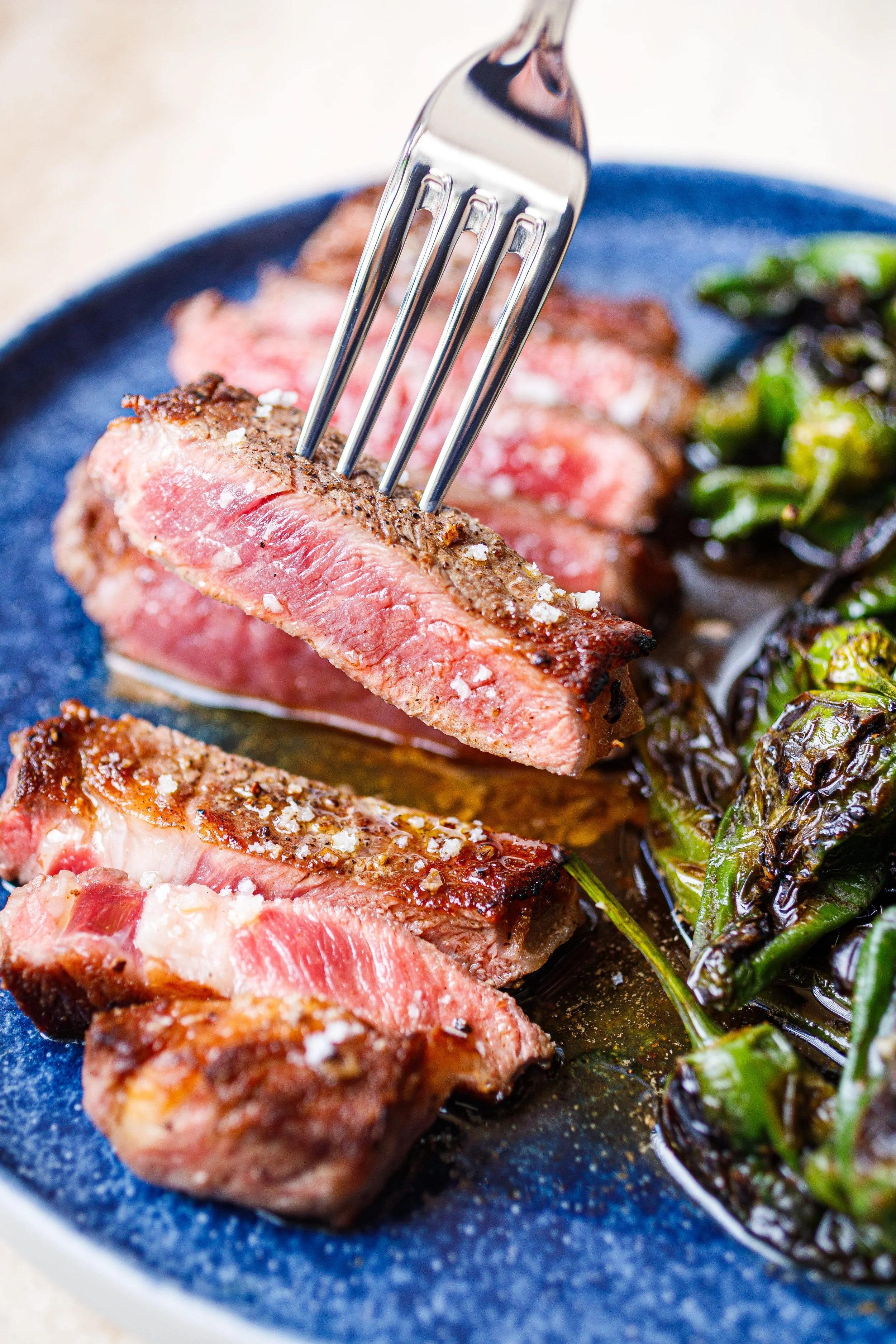 Close-up of medium-rare steak being picked up with a fork on a blue plate, served with roasted green vegetables.