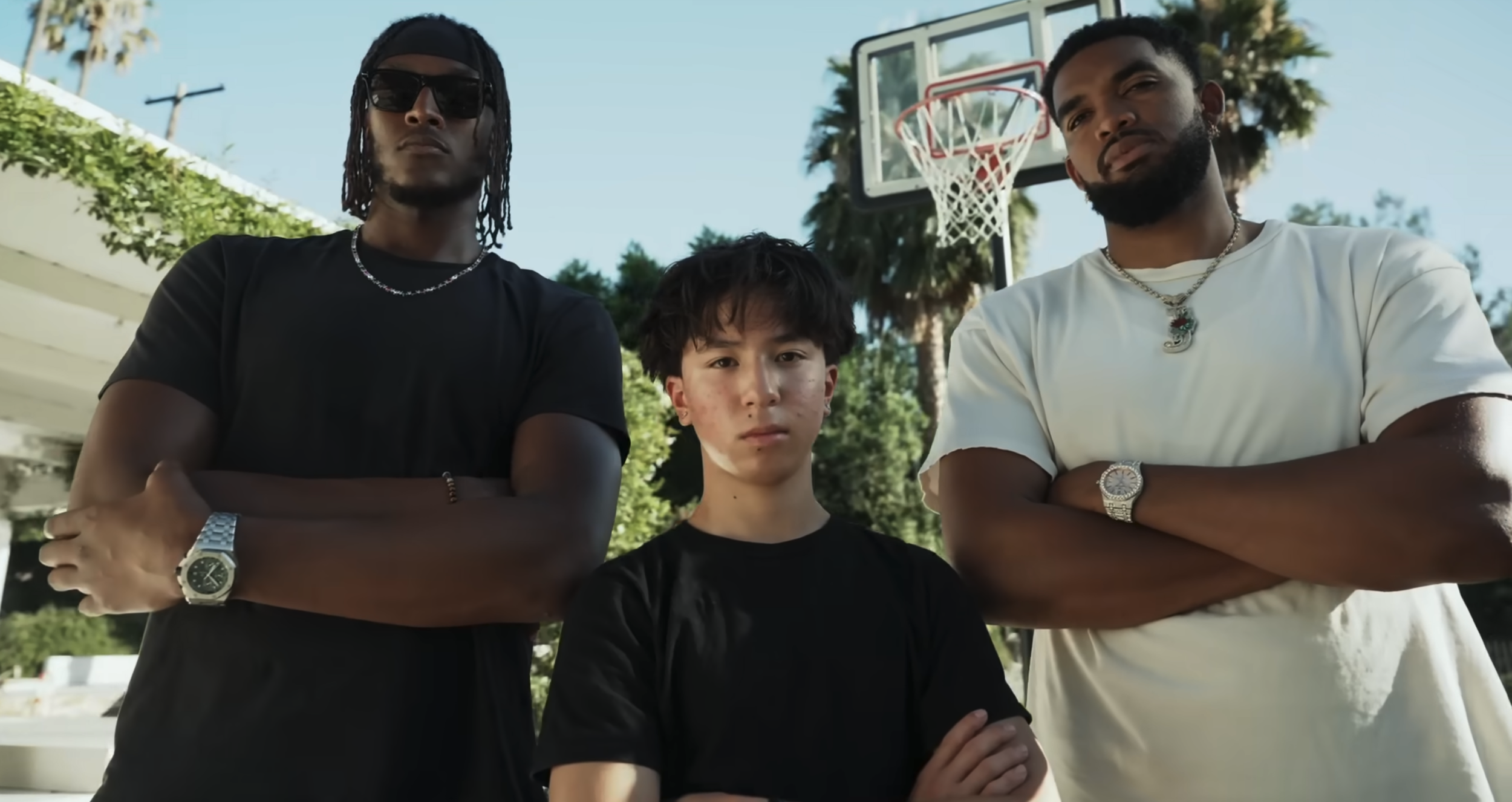 Three young men standing on an outdoor basketball court with a basketball hoop in the background. Two are adult men with arms crossed, and one is a teenage boy in the middle, looking serious. They are dressed casually with watches and jewelry.