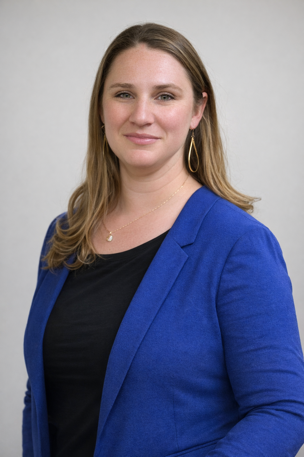 A professional woman with light skin, shoulder-length light brown hair, wearing a blue blazer over a black top, gold teardrop earrings, a delicate gold necklace with a small pendant, posing against a plain white background.