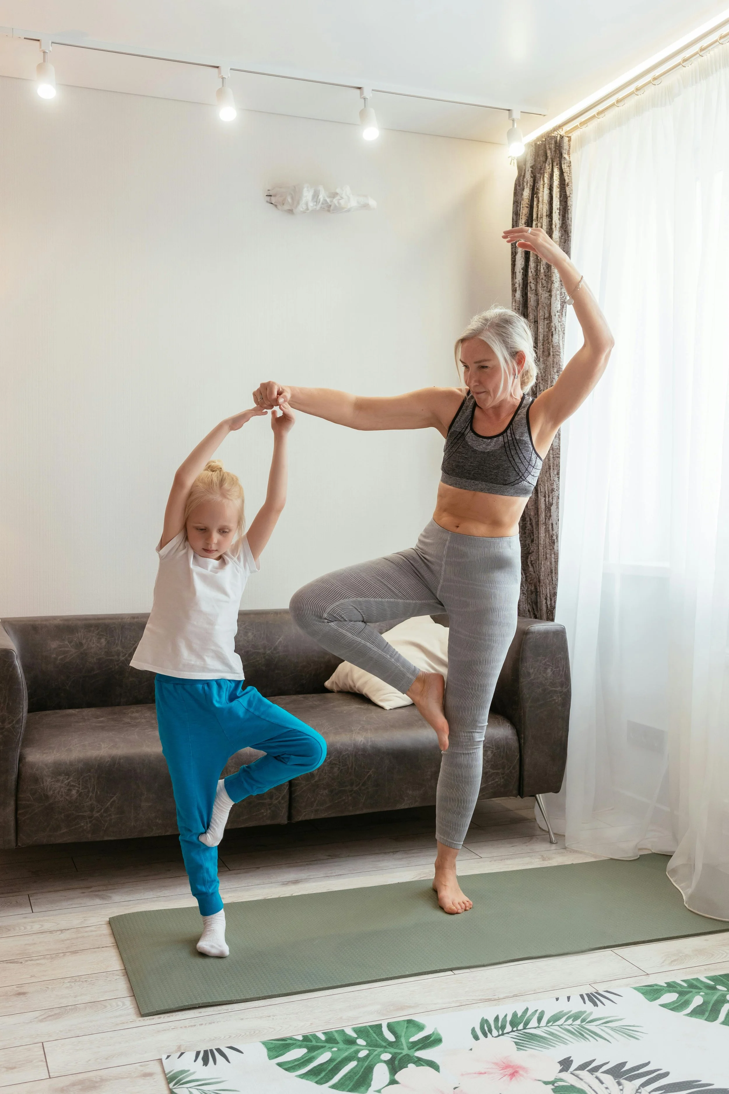 Une femme et une jeune fille pratiquant le yoga dans un salon, debout sur un tapis de yoga, en équilibre: la femme en position de l'arbre et la fille en posture de danse, tenant la main de la femme.