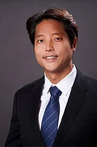 Professional headshot of a man in a black suit, white shirt, and blue tie, smiling against a gray background.
