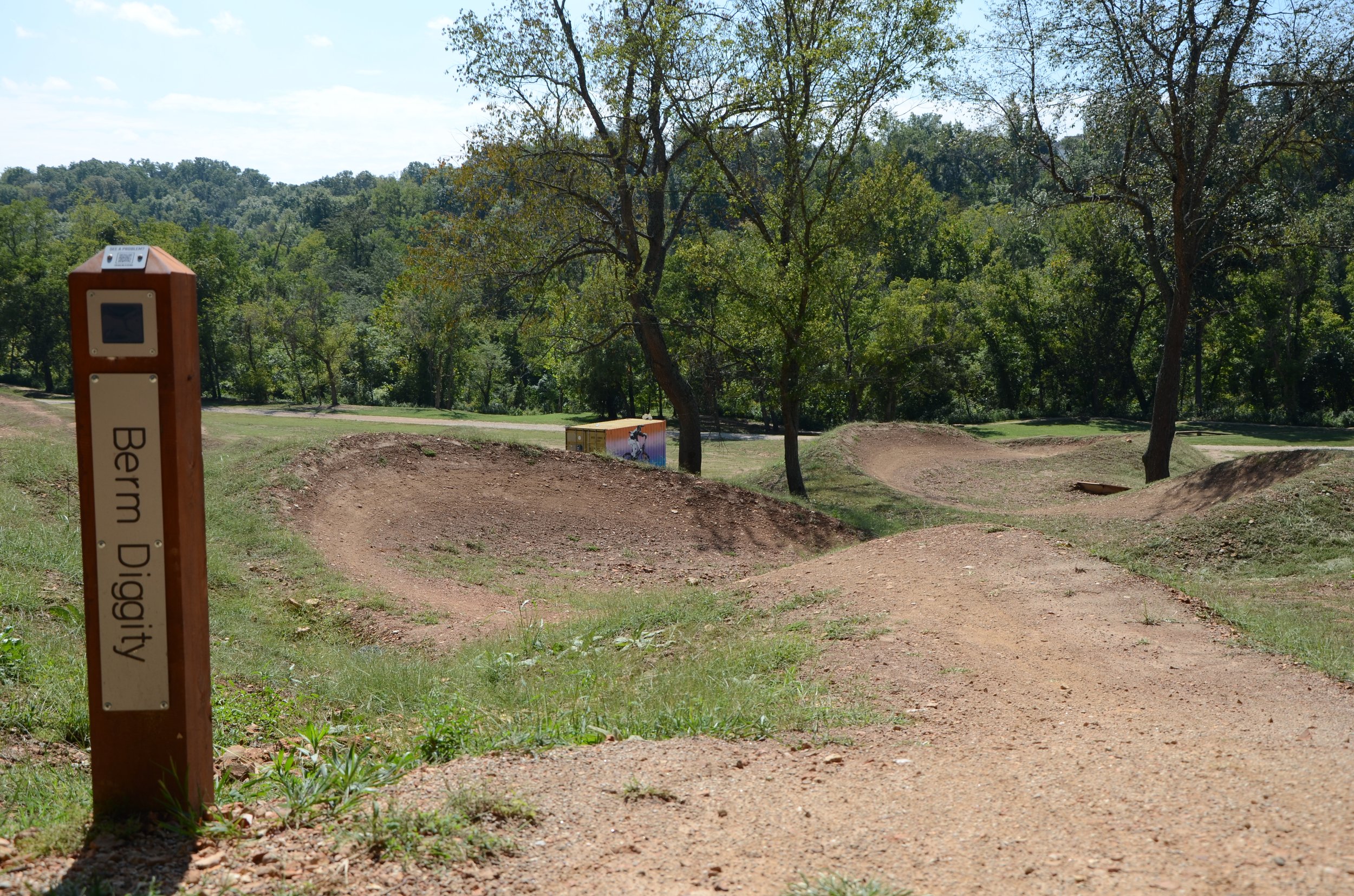 A picture of the trail sign for Berm Diggity in the Skills park Advanced Area and a view of the trail's big berms that weave near multiple tall trees in the Slaughter pen Trail system.