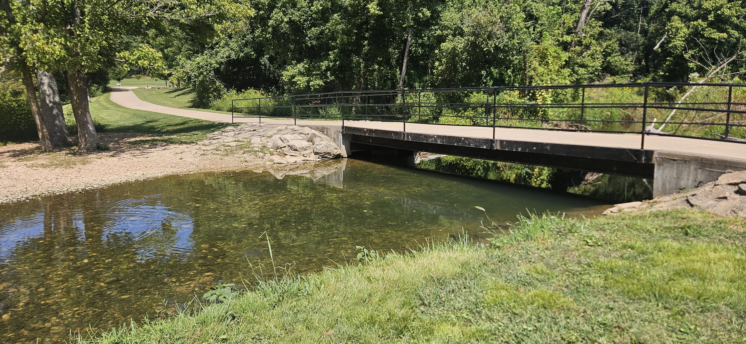 A small bridge across a shallow creek along the Razorback Greenway near the Skills Park in the Slaughter Pen Trail System. There is plenty of green grass along the creek for families and riders to relax.