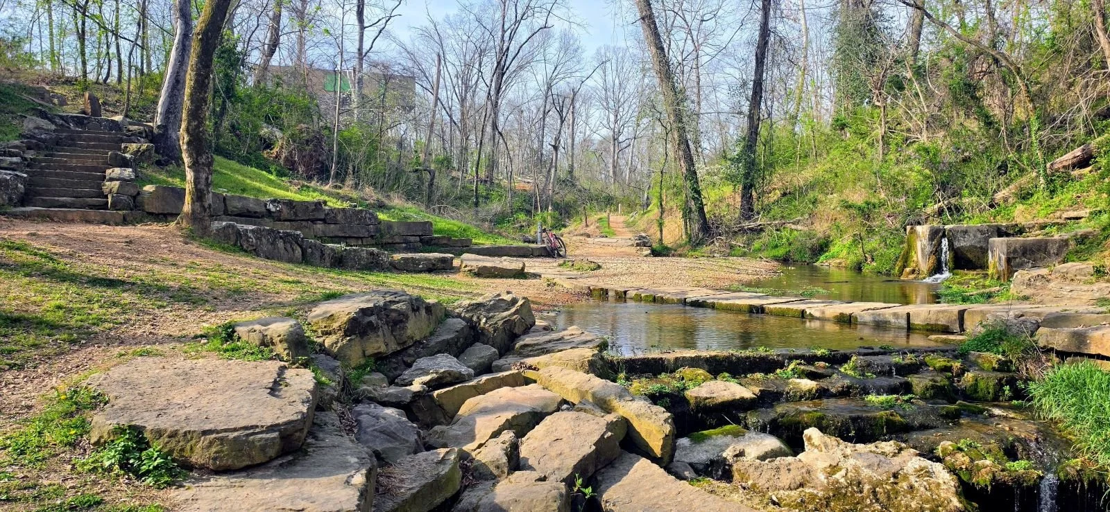 A peaceful creek with stepped waterfalls and rocks in a wooded park during spring, with bare trees and a bike resting on the trail.