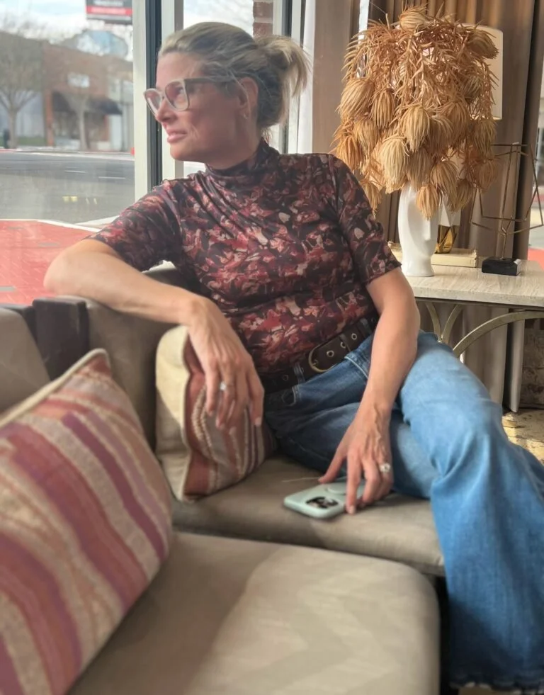 A woman with short blonde hair wearing an orange sleeveless top and beige pants sitting on a beige cushion, writing in a notebook. There is a large plant and abstract art with sun shadow on the wall.