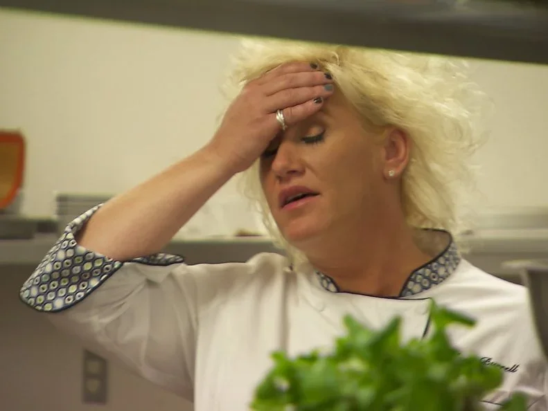 A woman with blonde hair, wearing a white shirt with patterned cuffs, is holding her forehead with her hand and appears stressed or upset, in a kitchen or food preparation area.