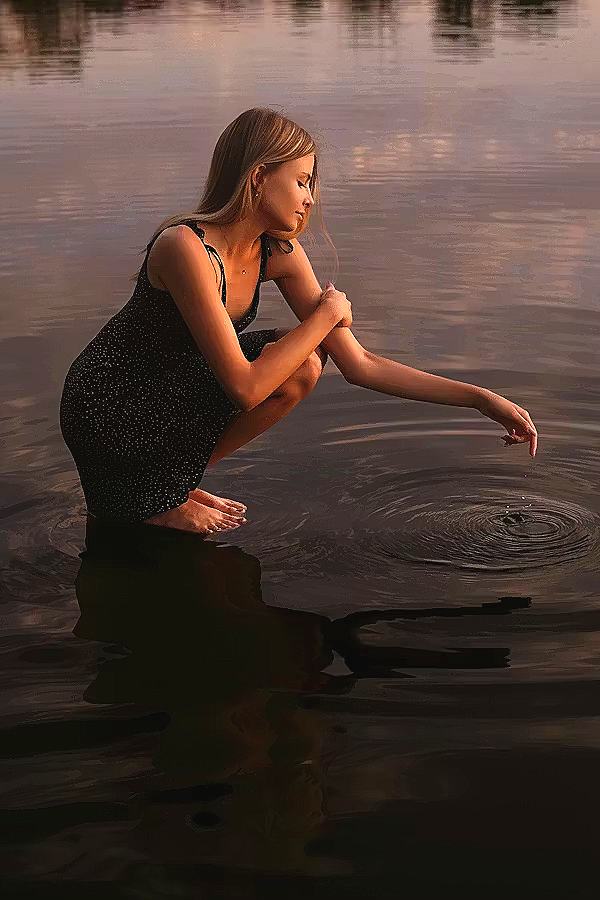A woman in a black dress squatting in shallow water, touching the surface with her right hand, during sunset reflection